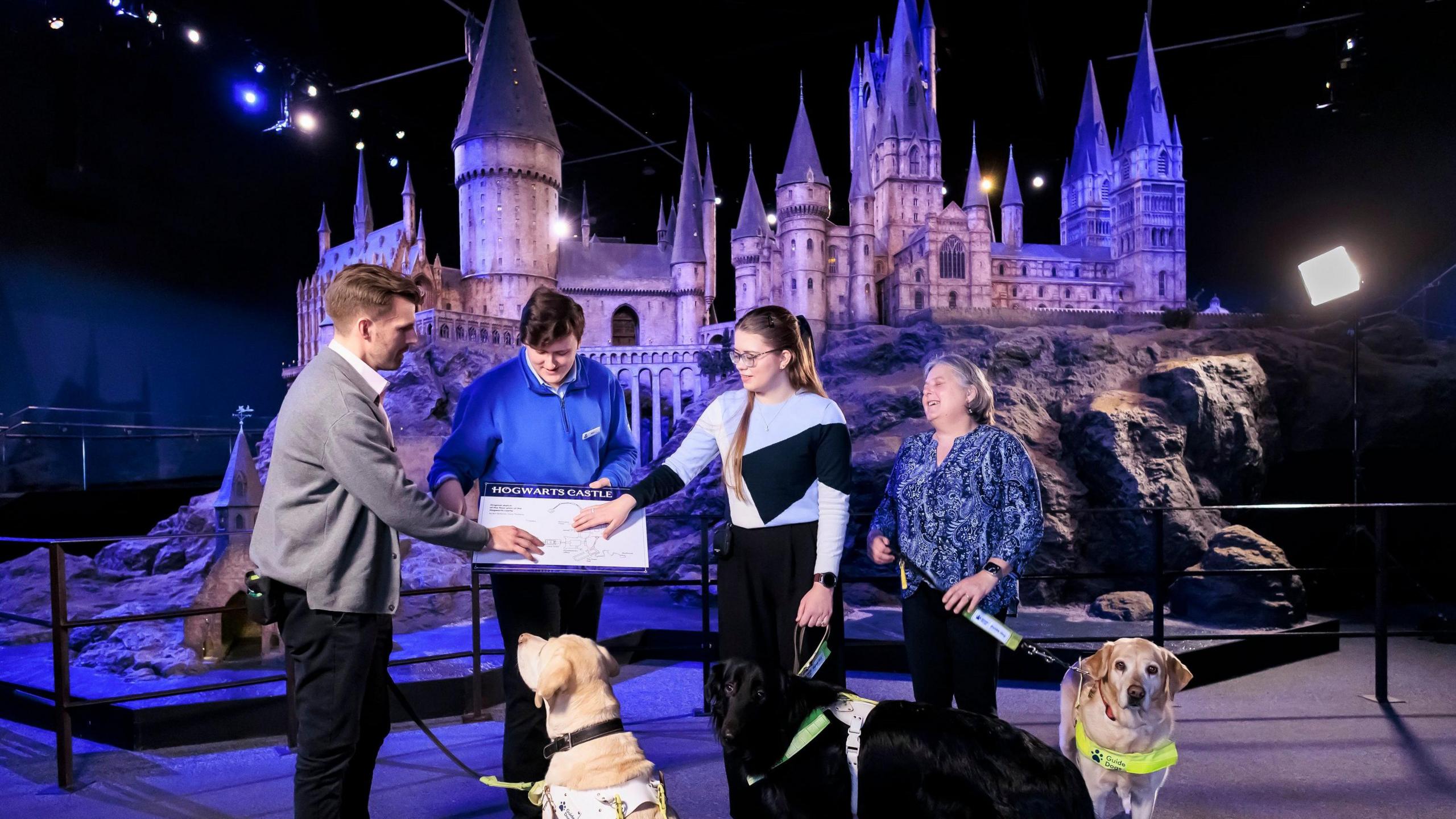 A man and woman are feeling a raised map of the Hogwarts miniature model. The actual model towers behind them. They are holding leads attached to guide dogs. Another woman is stood to the side also with a guide dog. A member of staff wearing a blue uniform is holding the raised map.