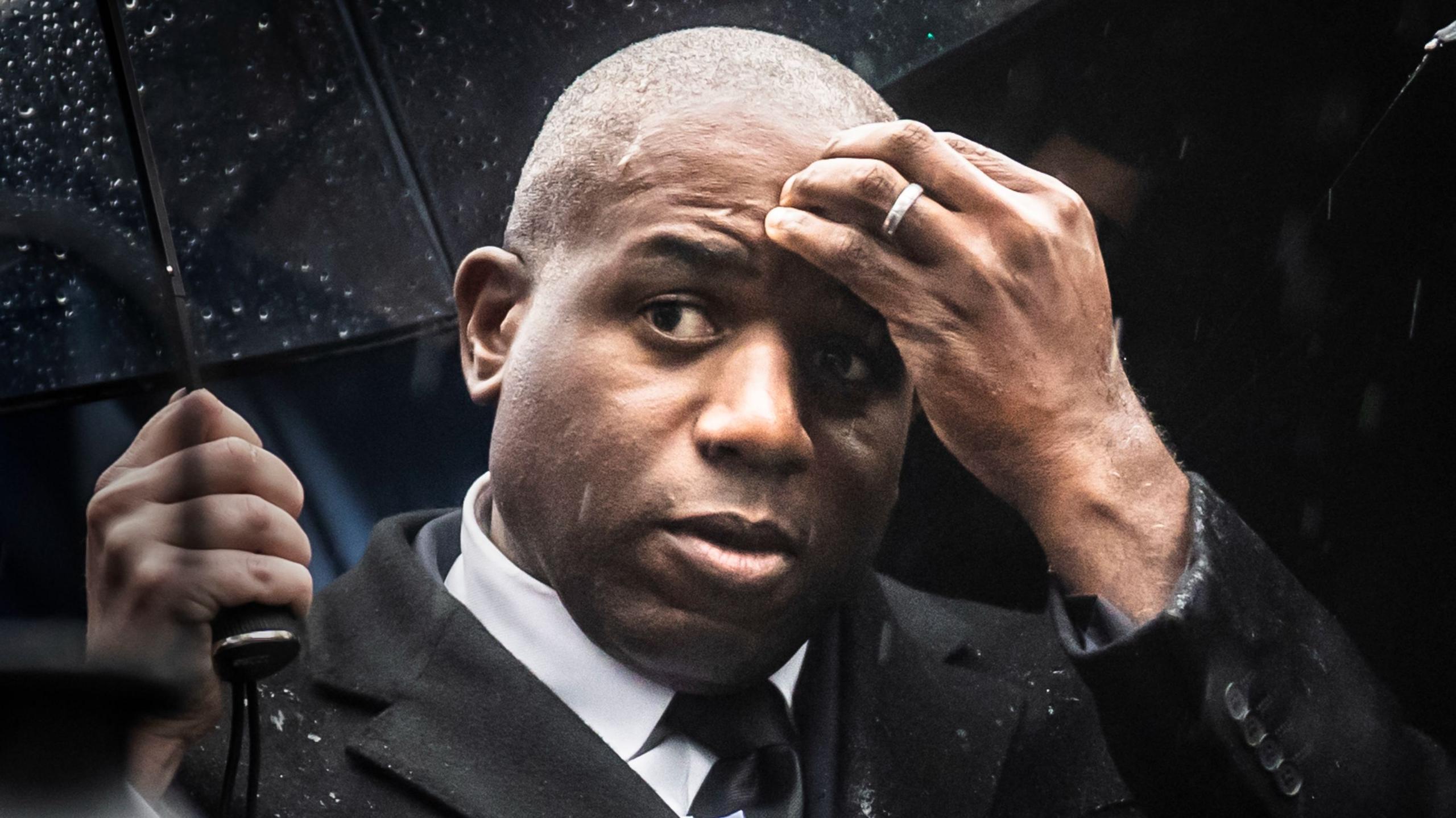 David Lammy wearing a black suit and black tie stands underneath a black umbrella. He is wearing an expression of concern and has his left hand raised to his forehead