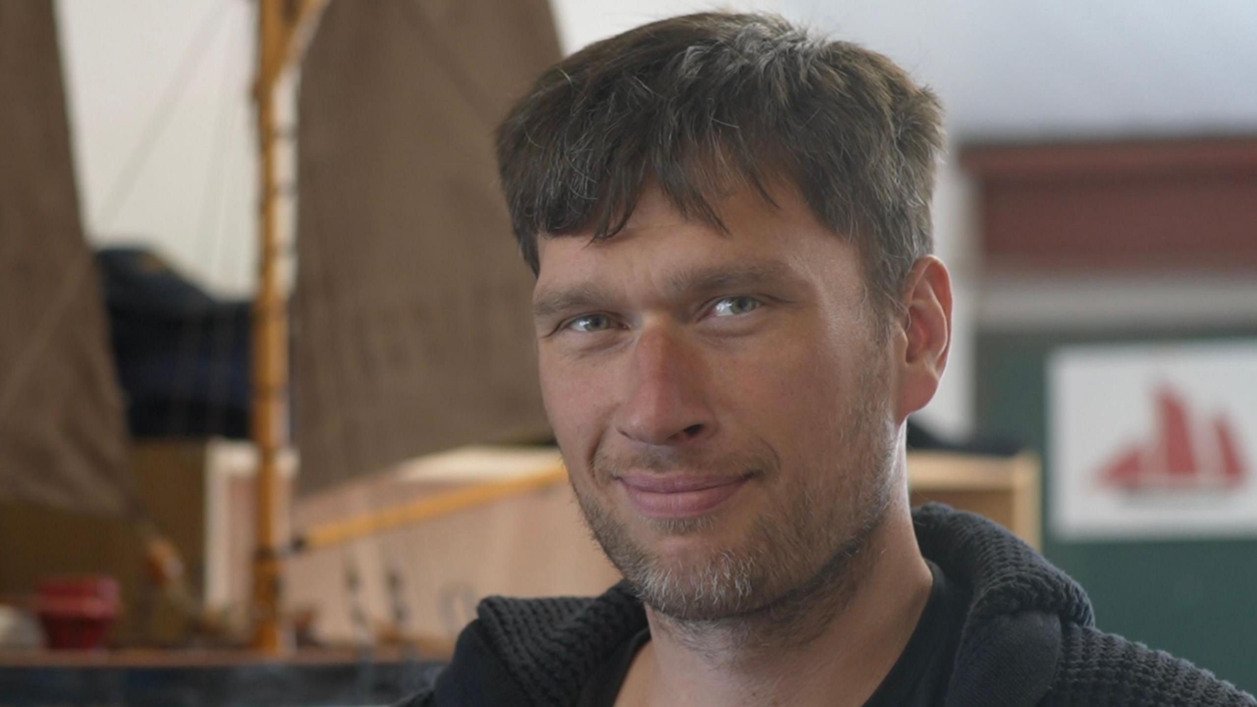 Karol Petryka is off centre to the right looking towards the camera. He has short brown hair and is wearing a black jacket. Behind him is a wooden replica of a boat.