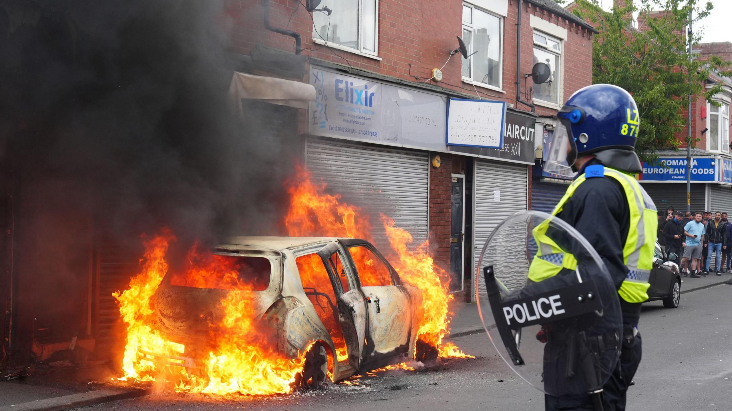 A car burns on Parliament Road, in Middlesbrough, during an anti-immigration protest. Standing, watching the flame, is a police officer in riot gear.