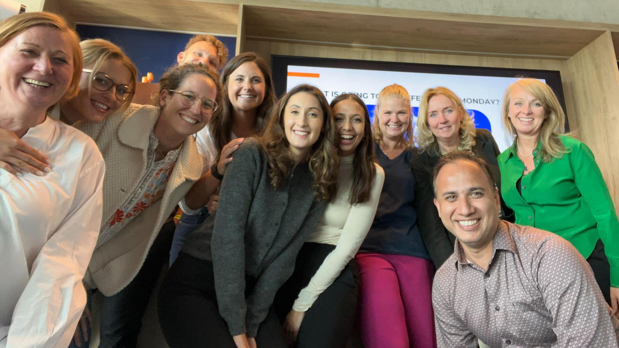 A group of 10 people (and another man at a lower height in front of them) standing together smiling in front of a tv screen that is almost entirely obscured.