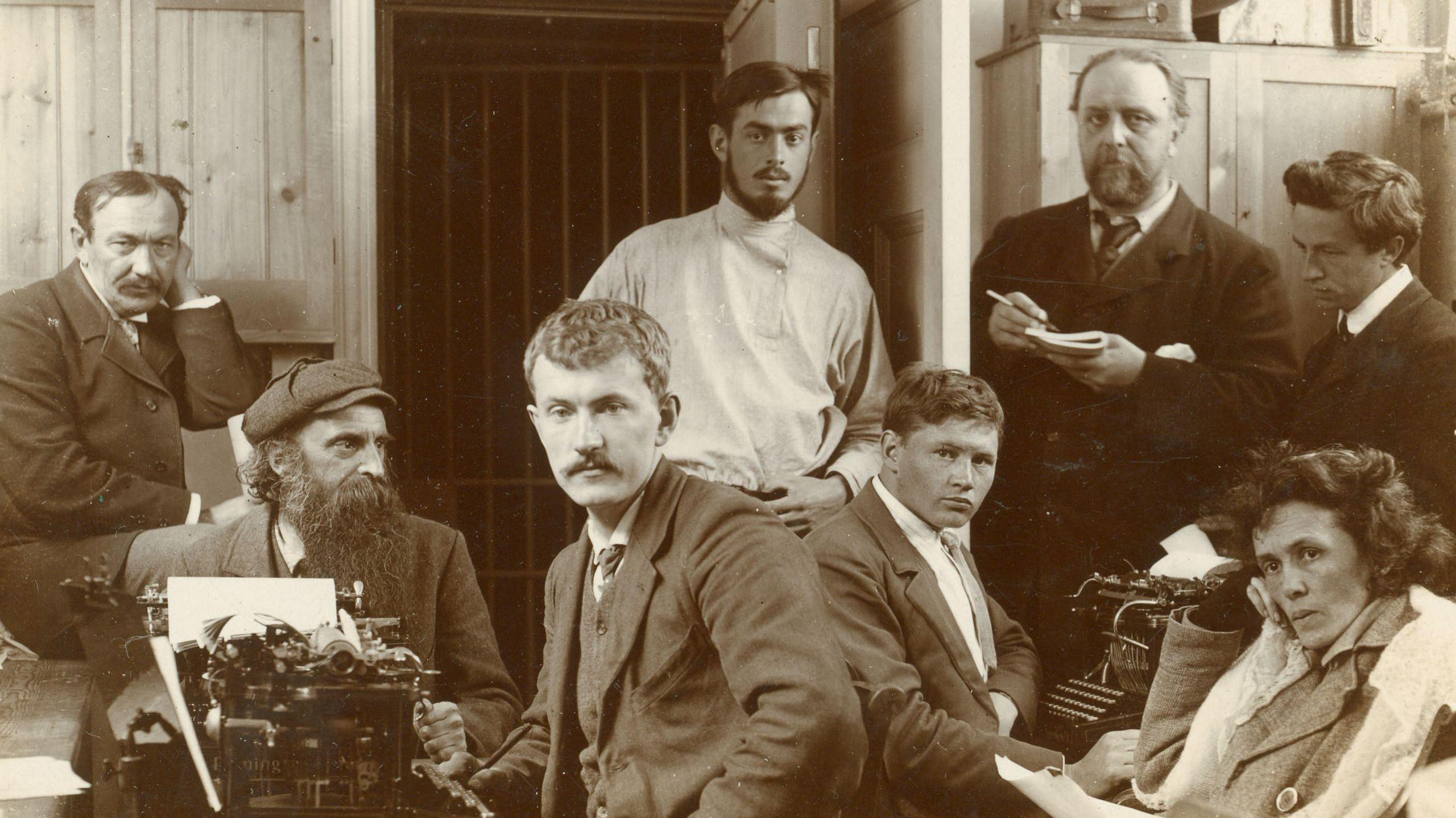 Sepia photograph of seven men and one woman sitting next to typewriters.