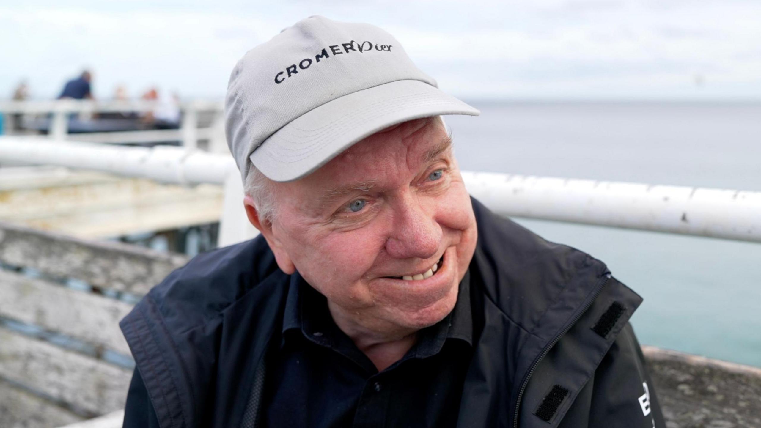 Nigel Hogg who is sitting on a bench on Cromer Pier. He is looking sideways and away from the camera. He is smiling and is wearing a black T-shirt with a black coat and a grey baseball cap which says, CROMER Pier, on it.