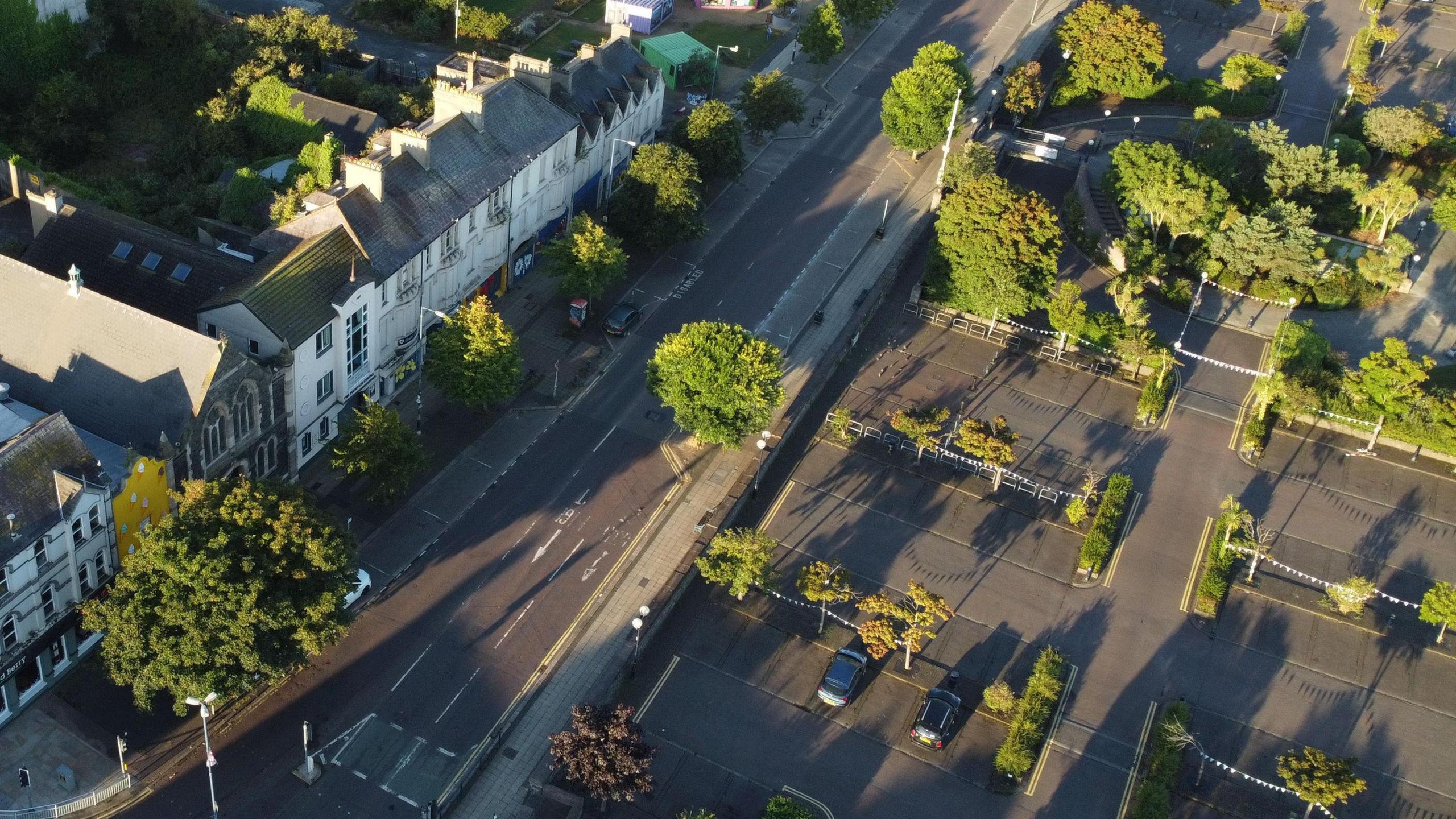 An aerial view of the Queen's Parade area of Bangor, County Down.
A number of buildings can be seen along a straight road opposite a large car park with various trees dotted throughout.