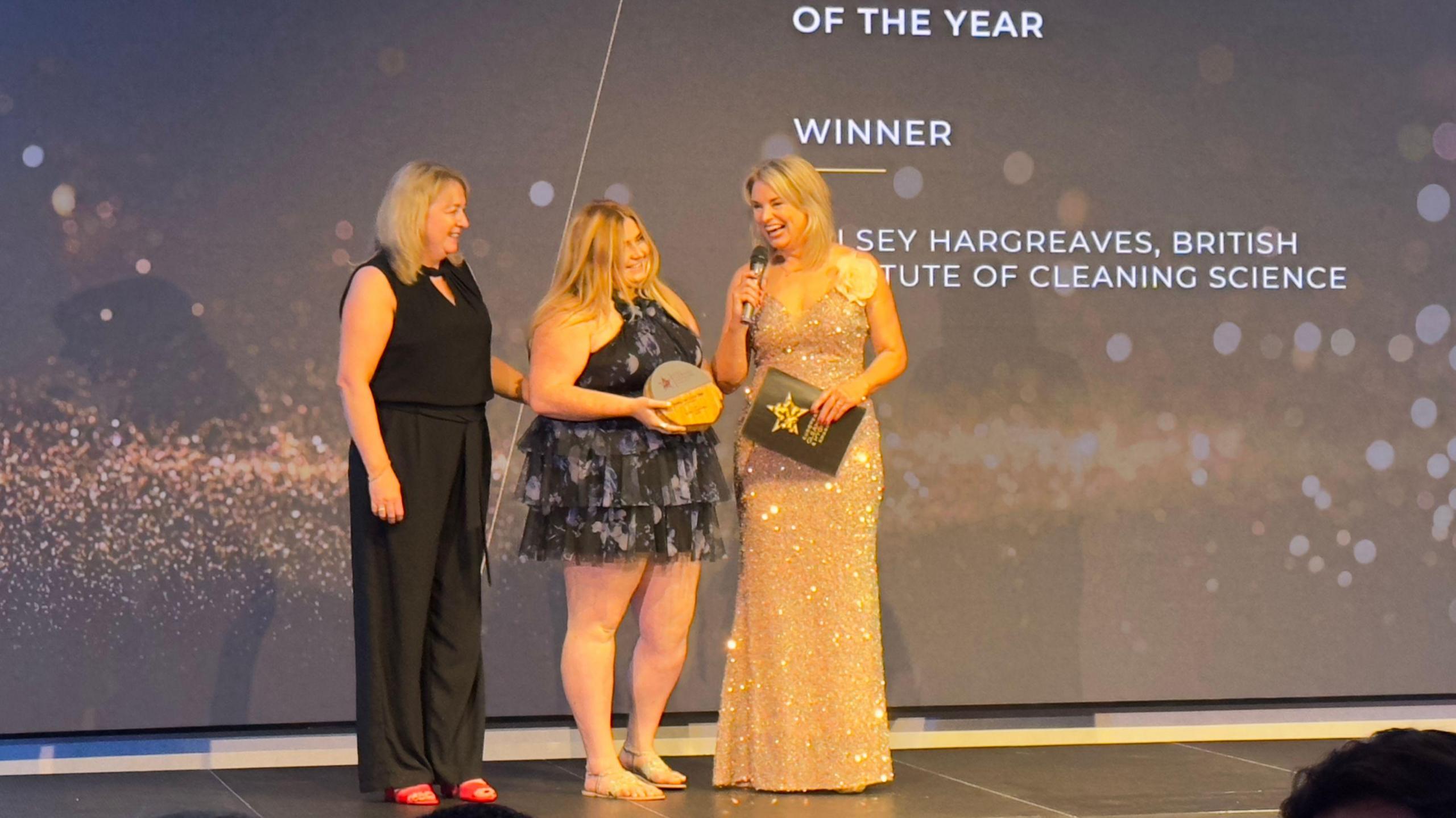 A woman in a black dress holds the arm of a blonde woman holding a circular trophy on a stage next to a blonde woman in a beige sparkly dress. 