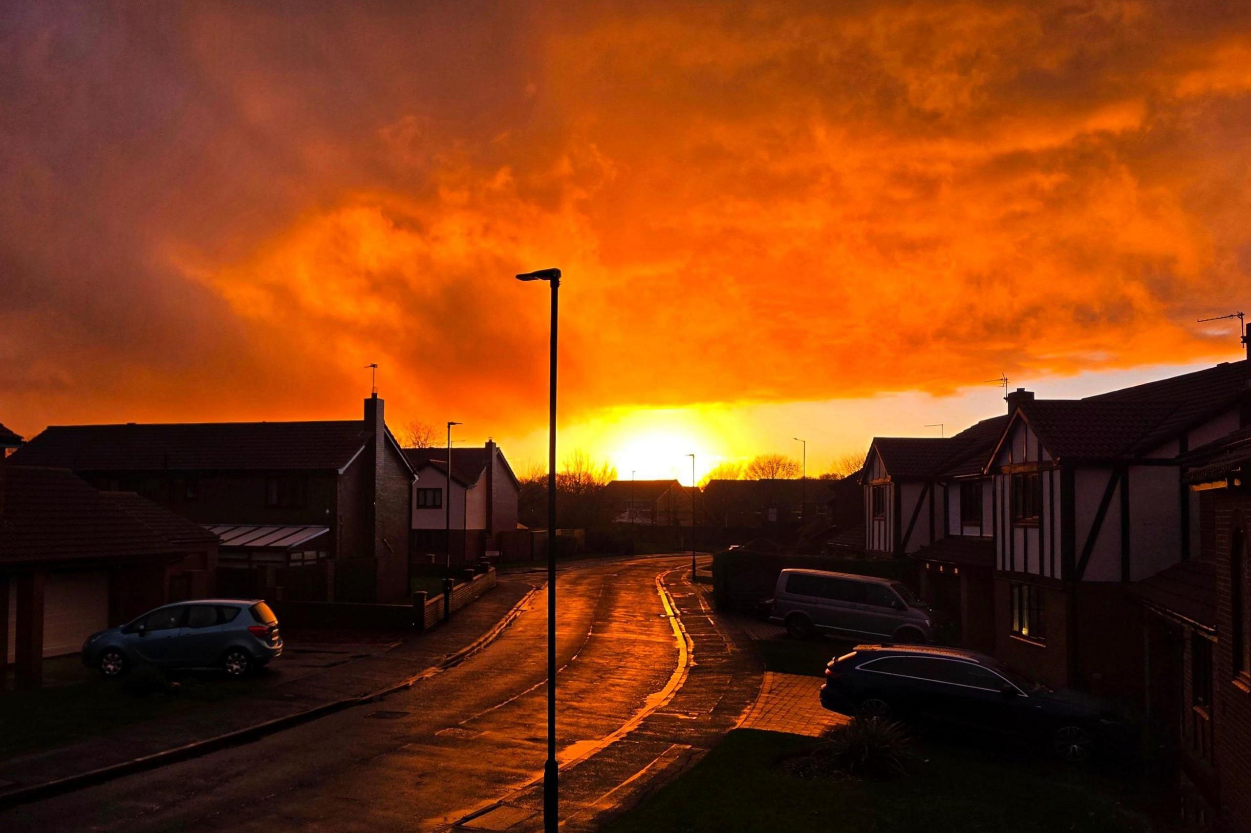 A residential street is illuminated as the sun sets. The clouds are a bright orange colour.