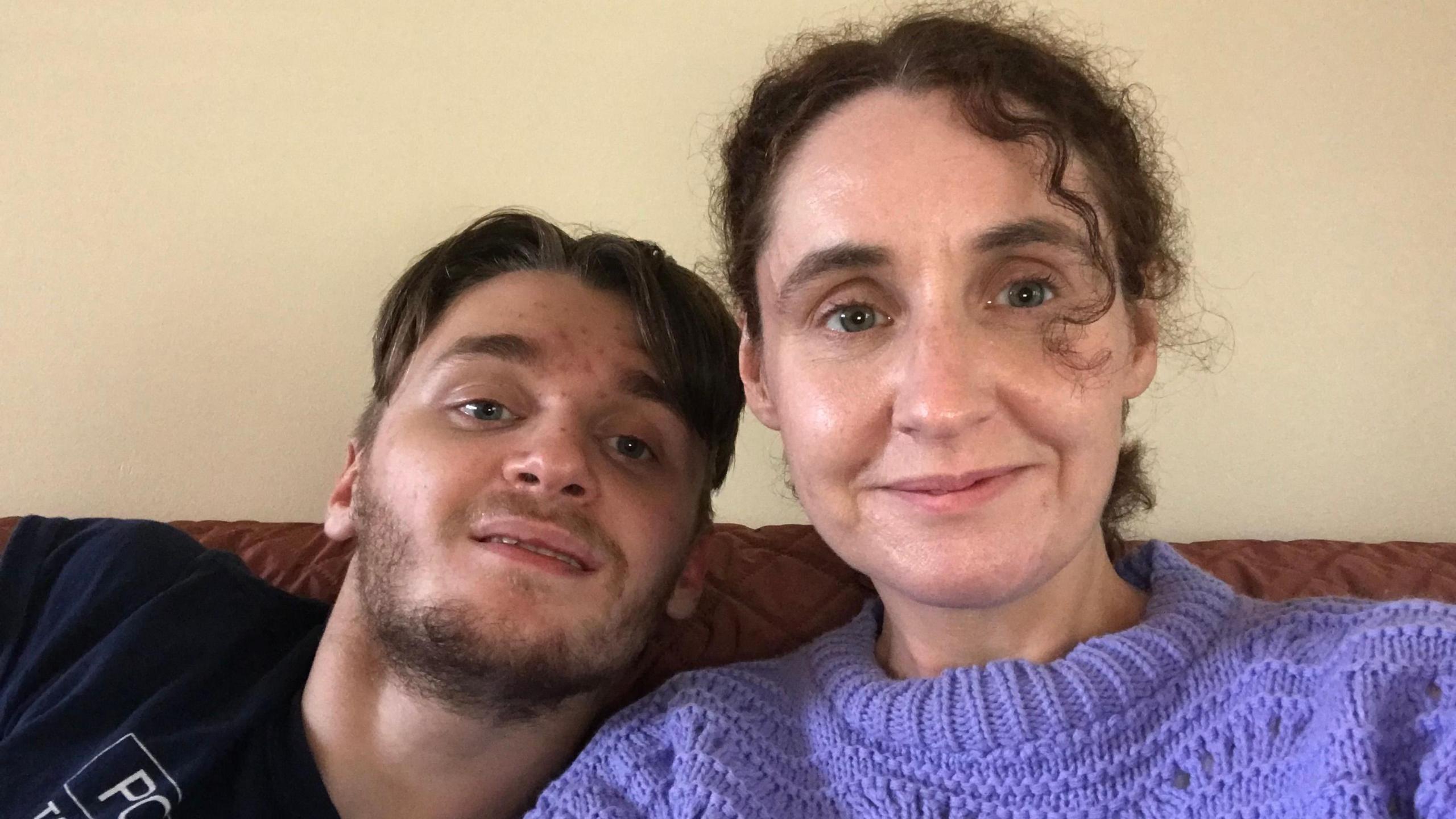 Jack and Dawn are sitting on a brown sofa, looking into camera. They are both smiling slightly. Jack has short hair and some facial hair. Dawn has curly brown hair tied back. She's wearing a light purple sweater and Jack is wearing a black t shirt with white writing on. 