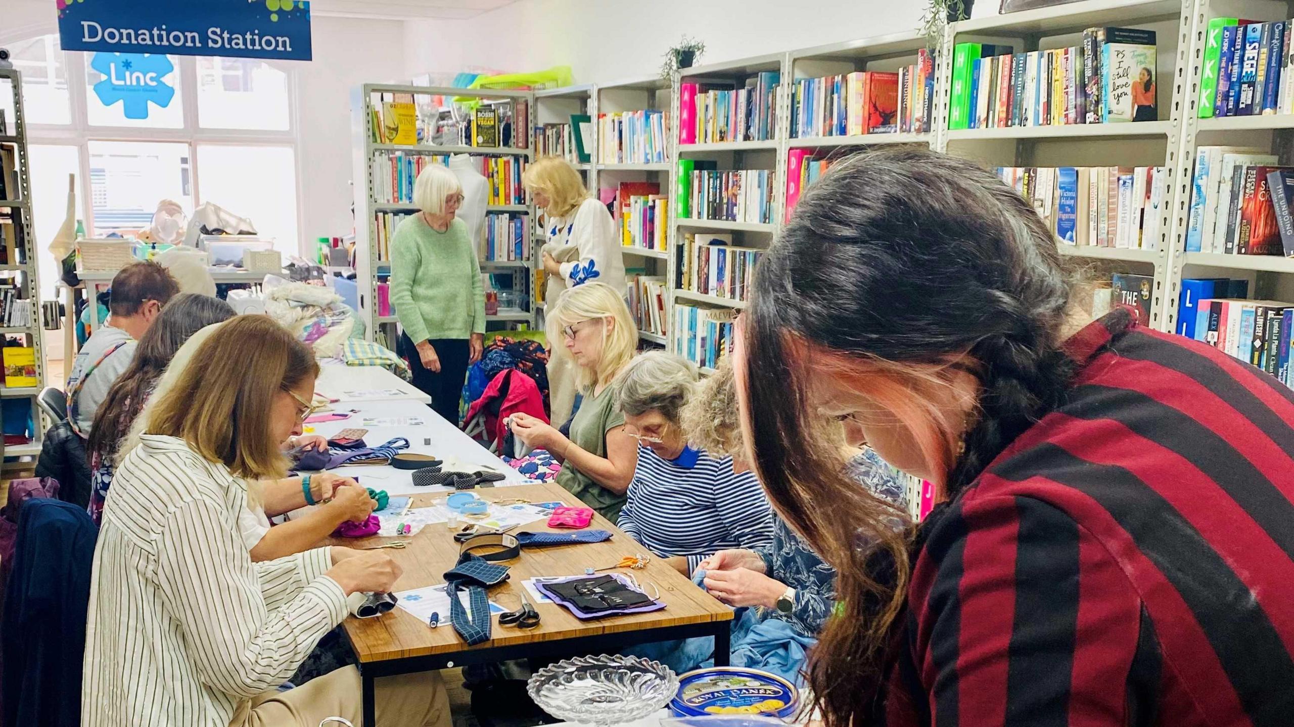 A group of people sitting around a large table in the Linc charity shop. They are sewing various items which you can see strewn across the table. There is a large sign saying 'donation station' in the background.