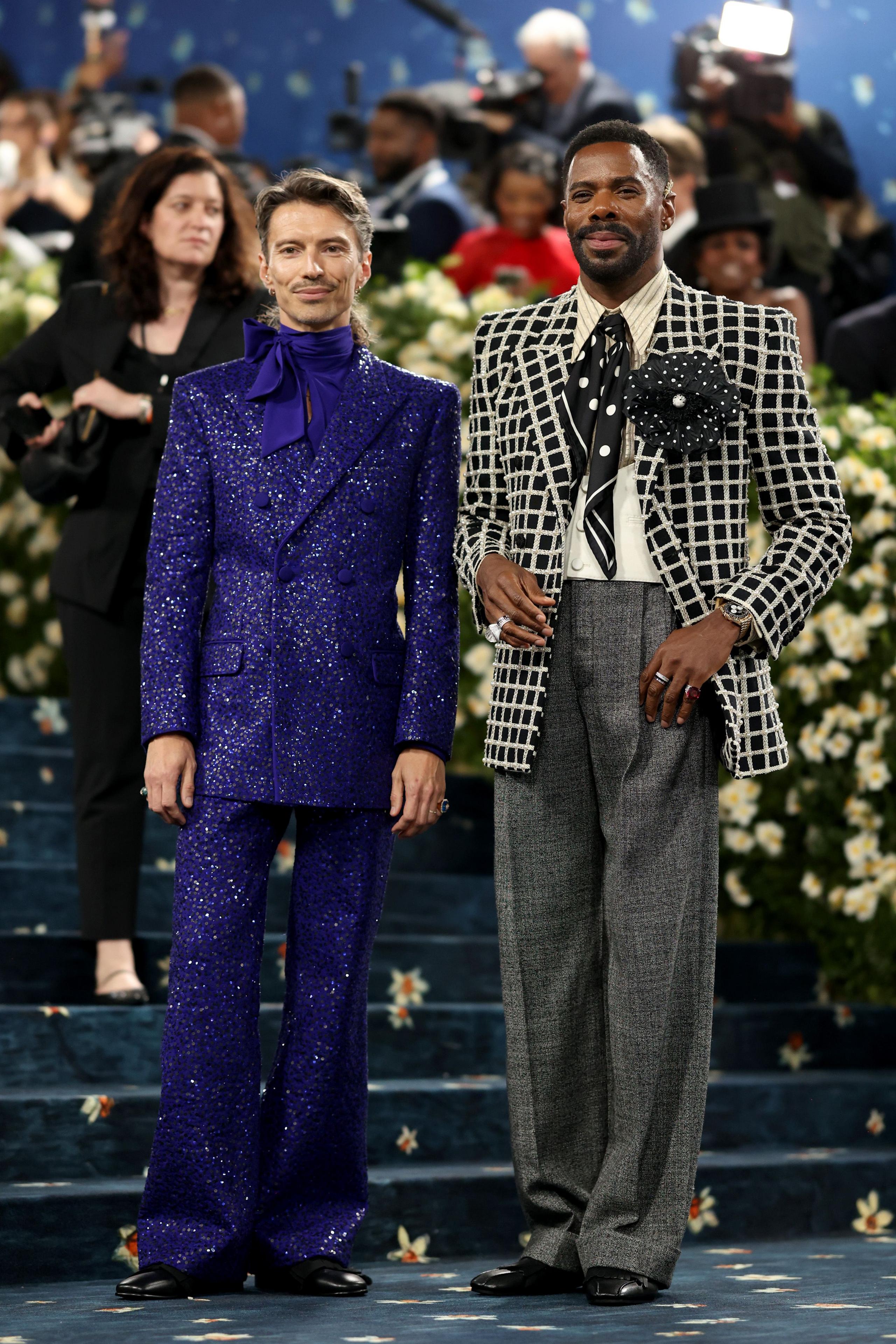 Colman Domingo stands with his husband Raul wearing a black and white jacket, a polka dot neckerchief and white shirt with a big black and white flower broach, while his husband wears a glittery tailored purple suit.