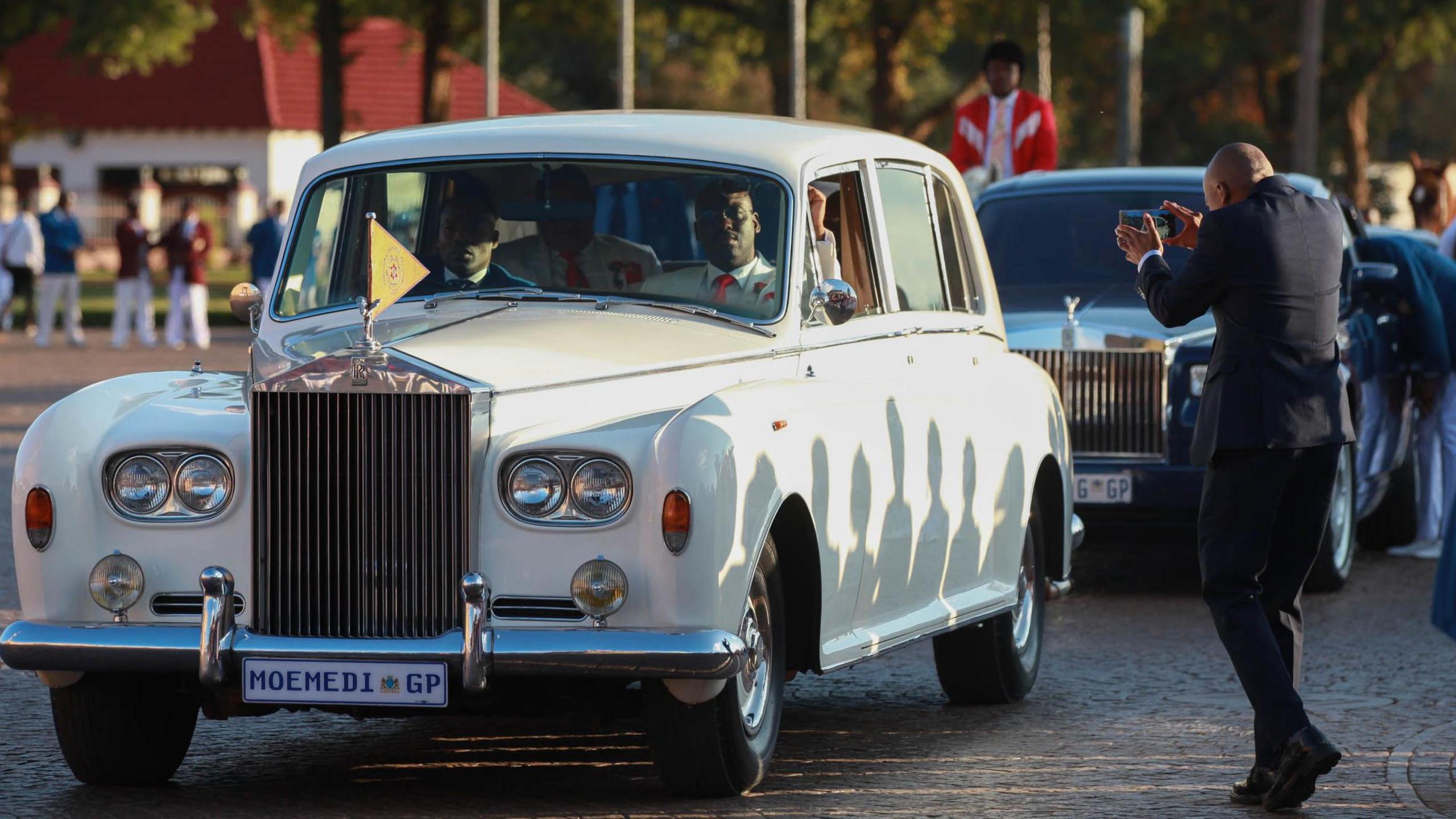 A convoy including a white Rolls Royce followed by a blue one arrives at the International Pentecost Holiness Church. A man in a black suit takes photos of them. Behind the second Rolls Royce, a man on horseback in a red blazer, can be seen.