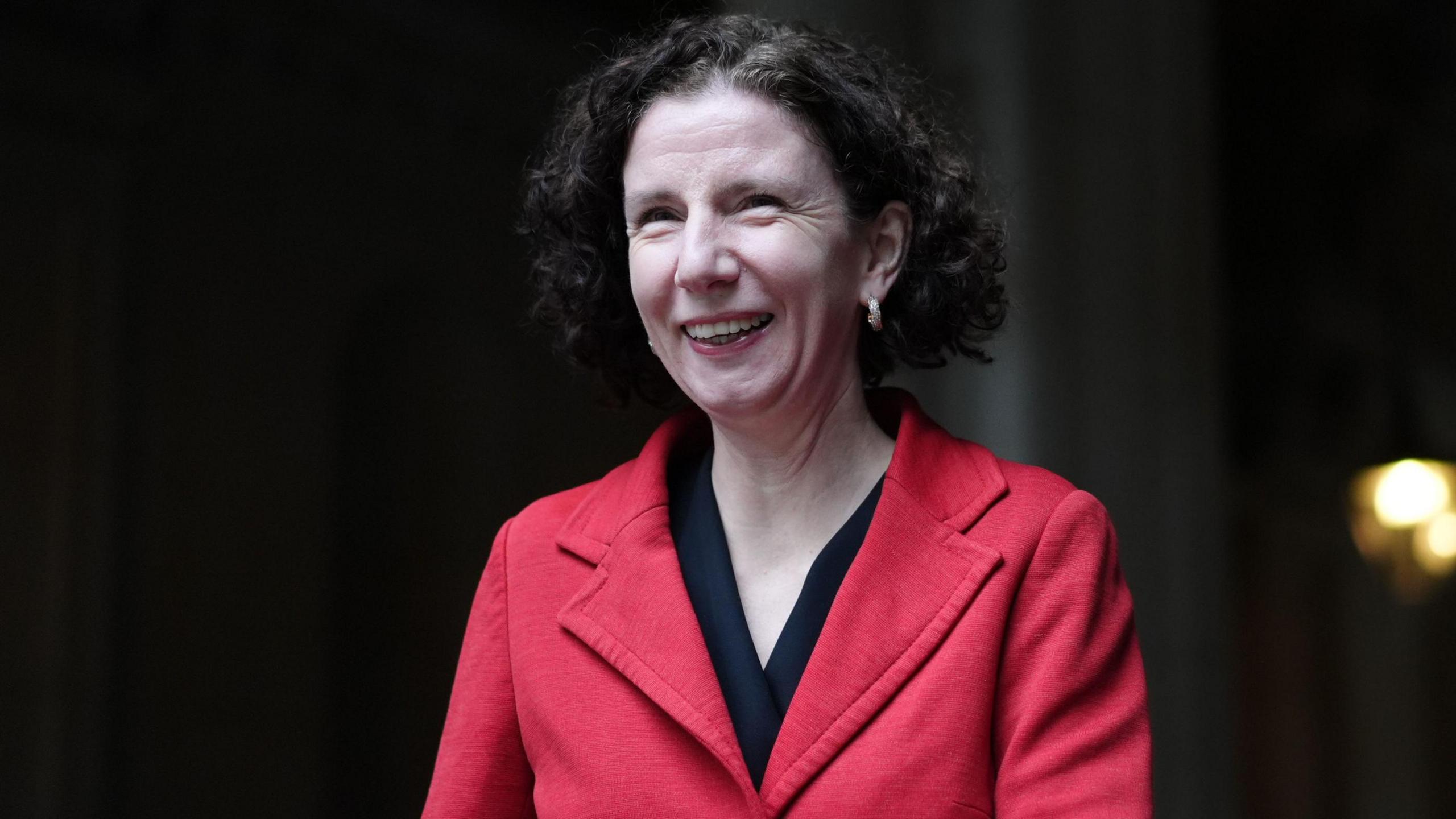 Anneliese Dodds has medium length brown curly hair and is wearing a red jacket over a black top.