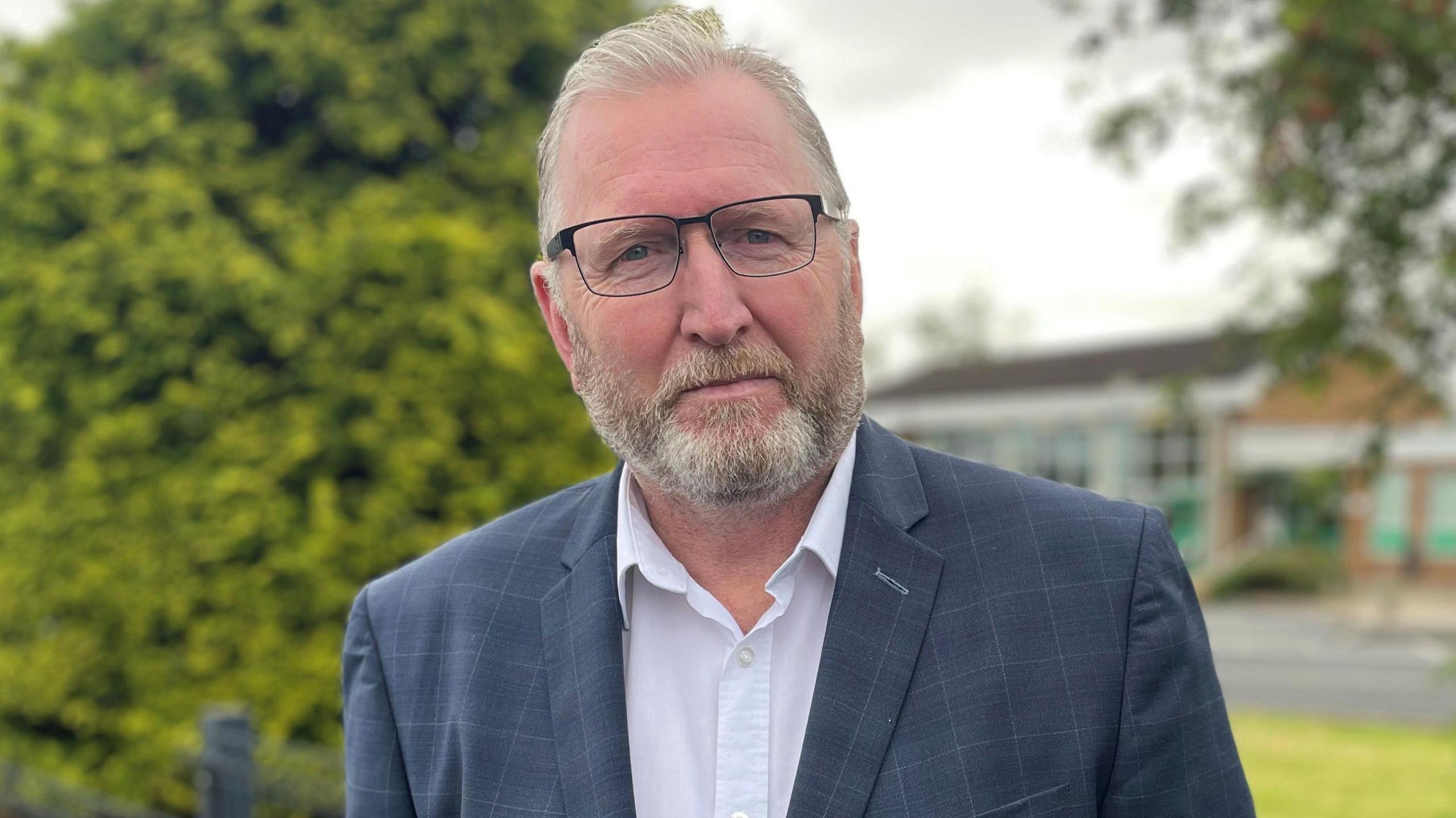 Doug Beattie has short white hair and a beard. He is smiling at the camera. He is wearing a pair of wire framed square glasses, a blue plaid suit jacket and a white collared shirt.
