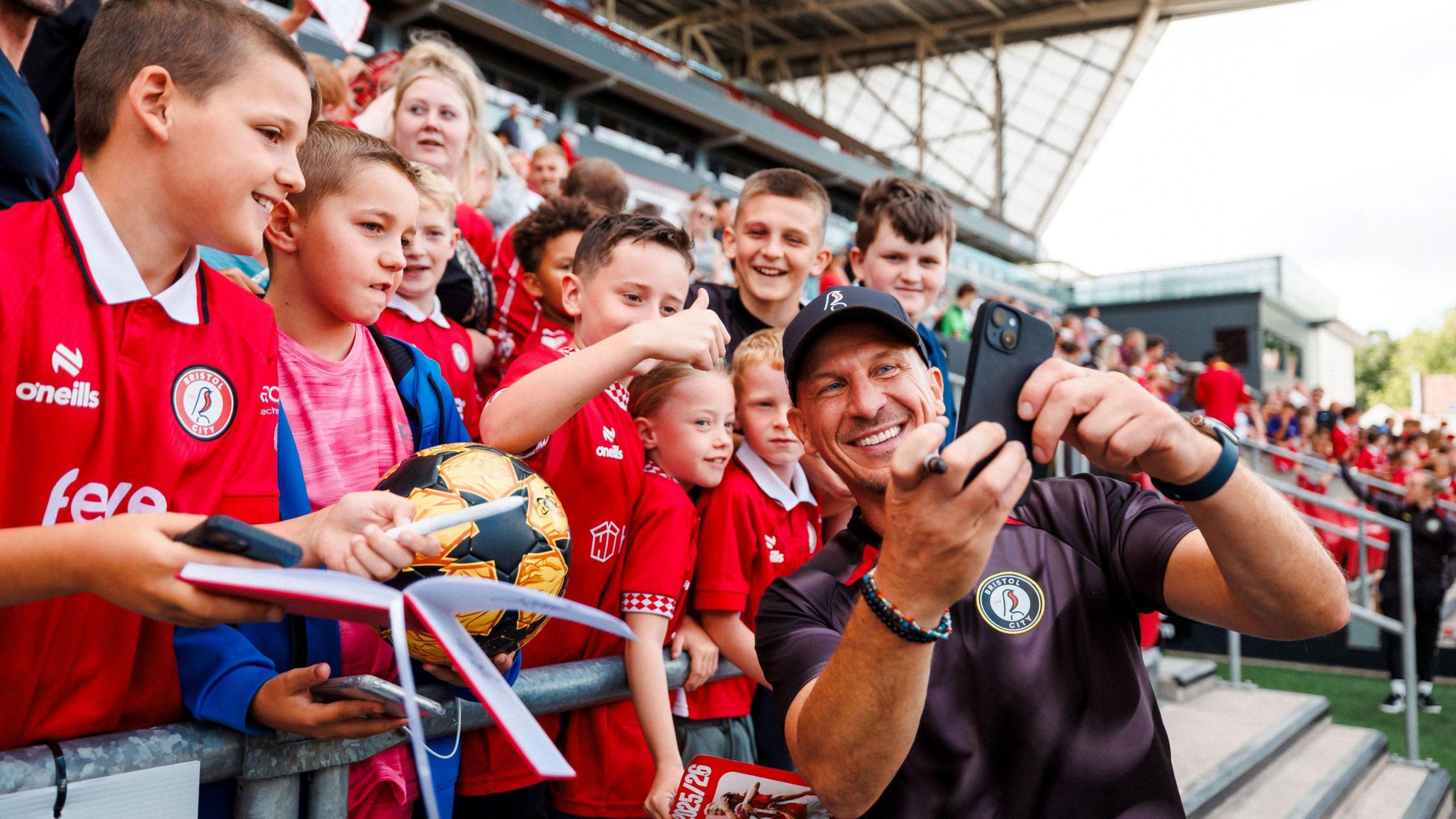 Bristol City manager Gerhard Struber smiles as he takes a selfie with young fans during the club's open day at Ashton Gate stadium. He is wearing a cap and a black training top, and the young fans are mostly boys wearing red football tops.