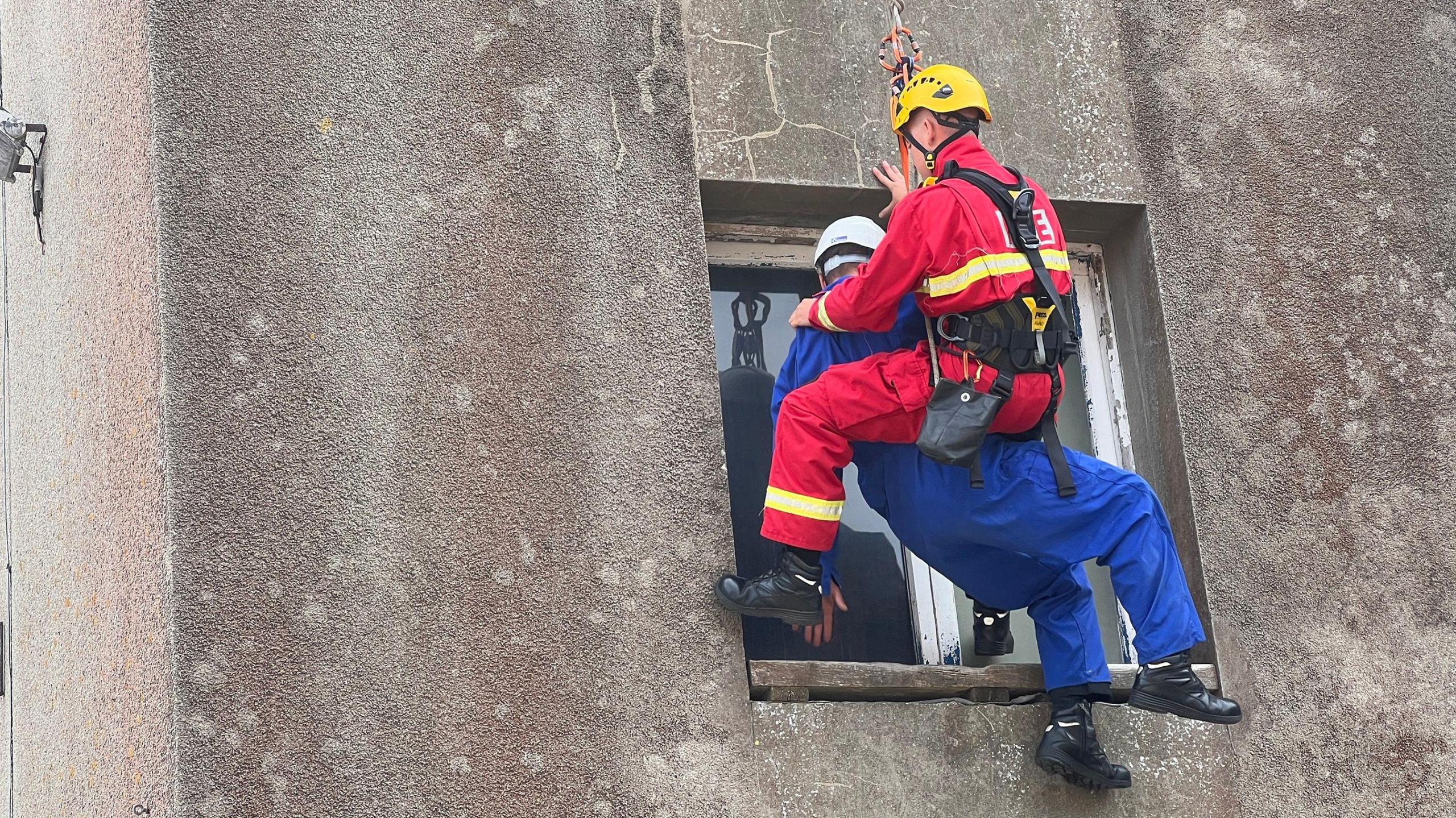 Logan is in a harness infront of a window as he is being lowered down. He is holding onto the casualty. Logan is in a red jumpsuit while the casualty is in a blue one. 