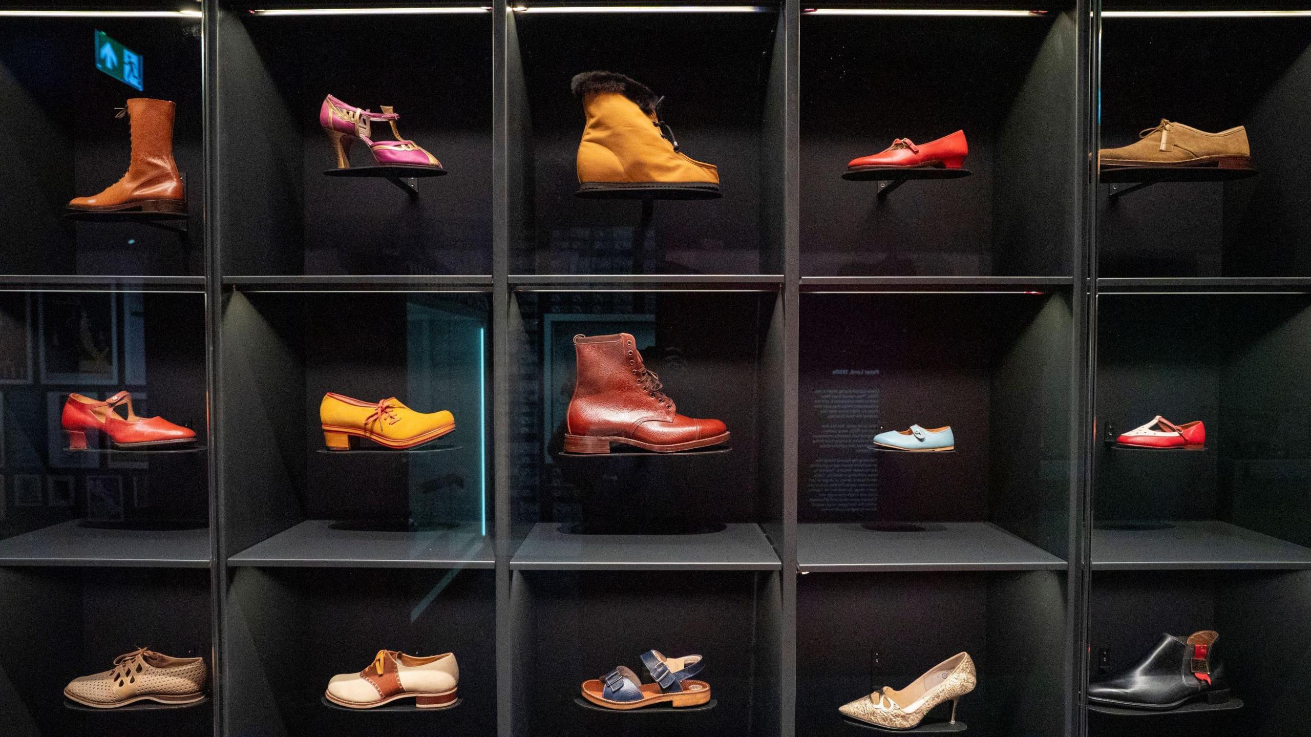 A display cabinet made of dark materials at the Clarks Museum in Somerset. It is divided into smaller boxes and in each box is a single shoe, all of them different designs. They include boots, high-heeled shoes, chunky vintage shoes and children's shoes