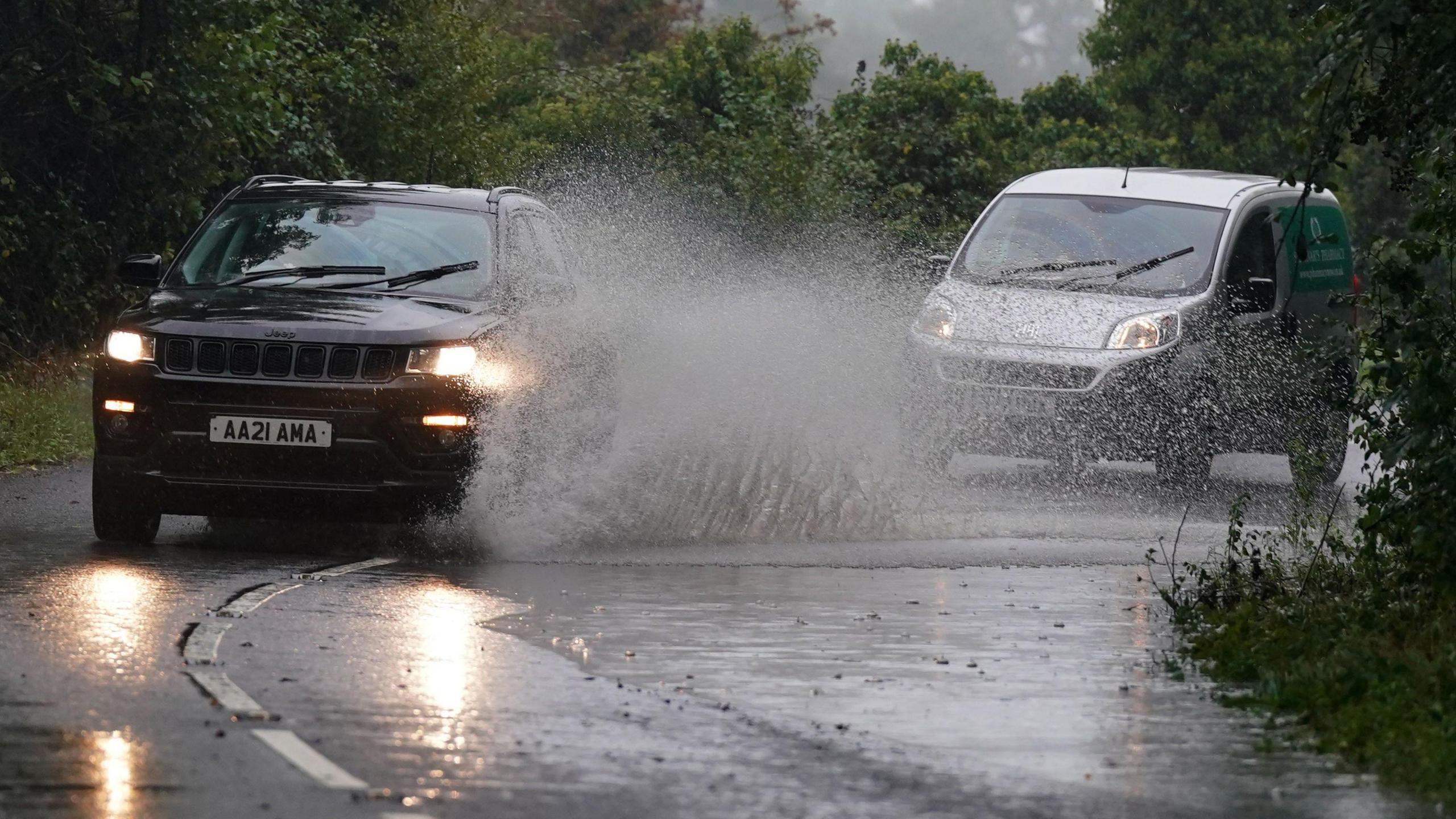 Cars drive through surface water in Willersey village, Gloucestershire. There is a large splash of water that reaches the car's roof. 