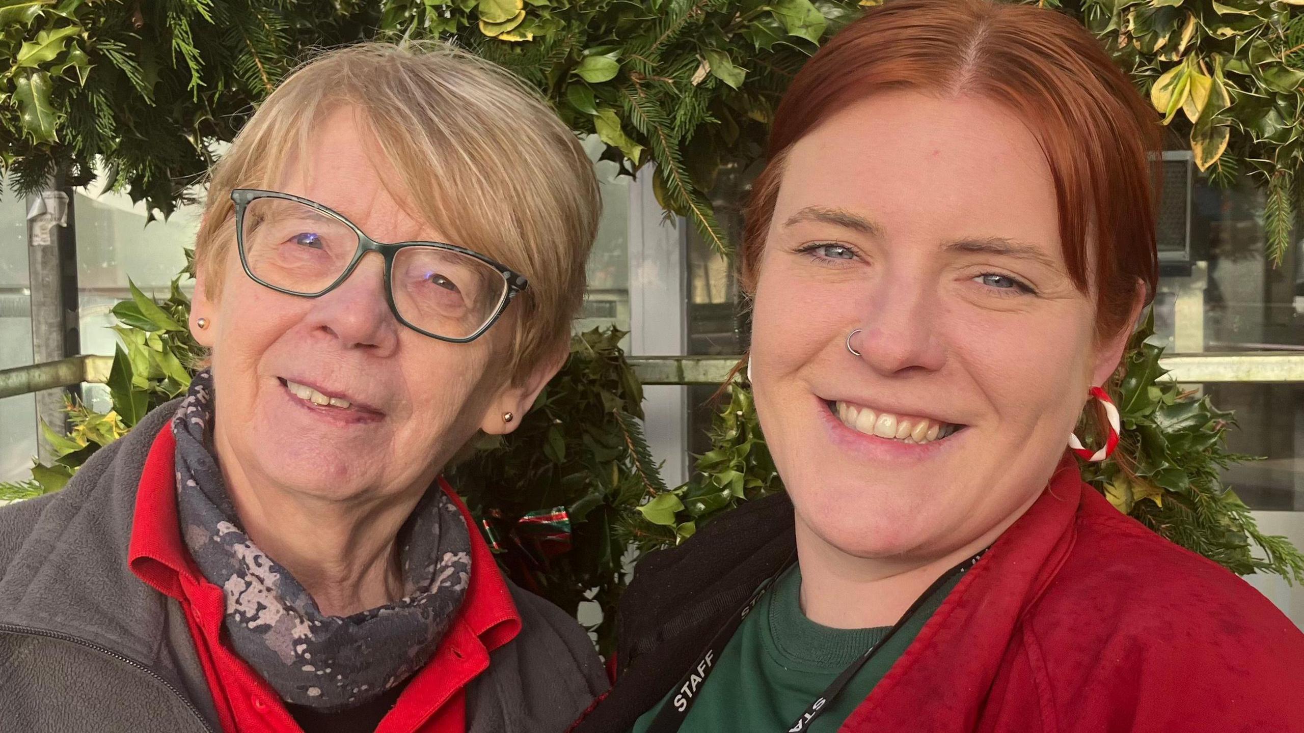 Both women are smiling into the camera.  They are both wrapped up warmly as they stand outside.  The woman on the left is wearing glasses; she has a grey scarf under a red t shirt which is topped off by a grey fleece jacket.  The woman beside her is wearing a red fleece jacket under a green t shirt.