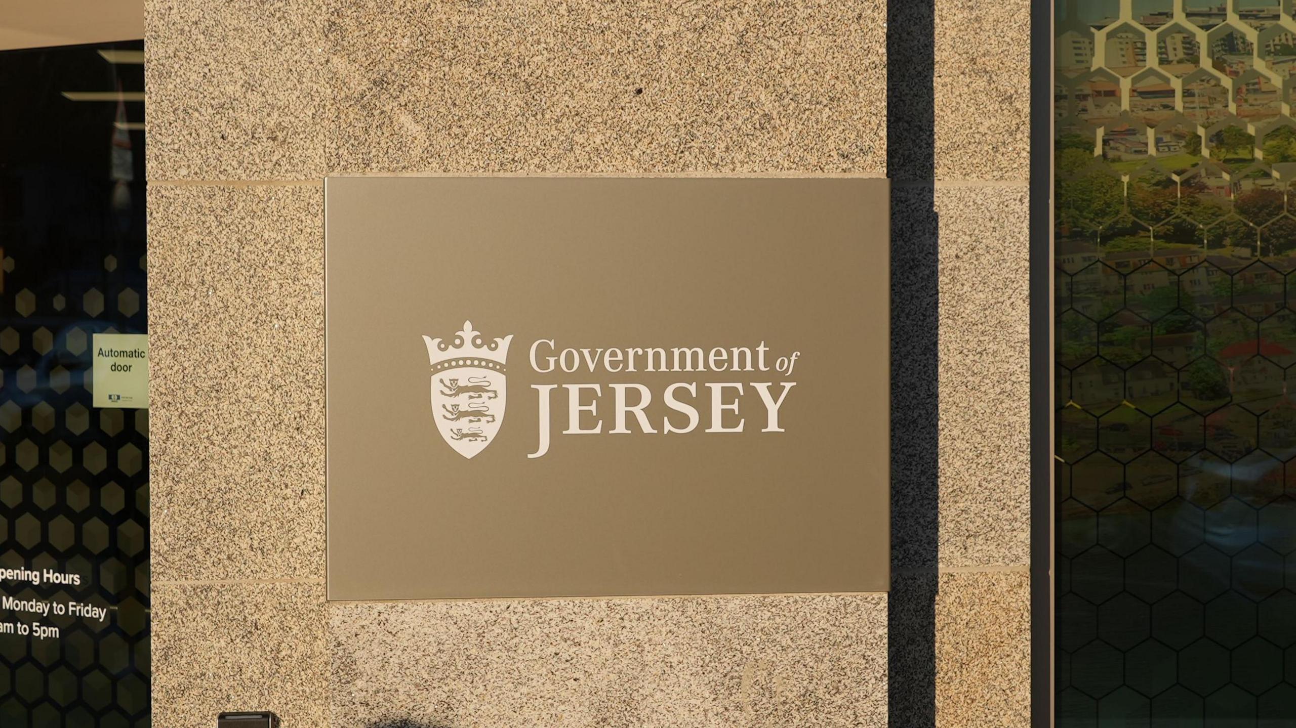 A gold and white coloured Government of Jersey sign on the side of a building. An automatic door is to the left of the sign.