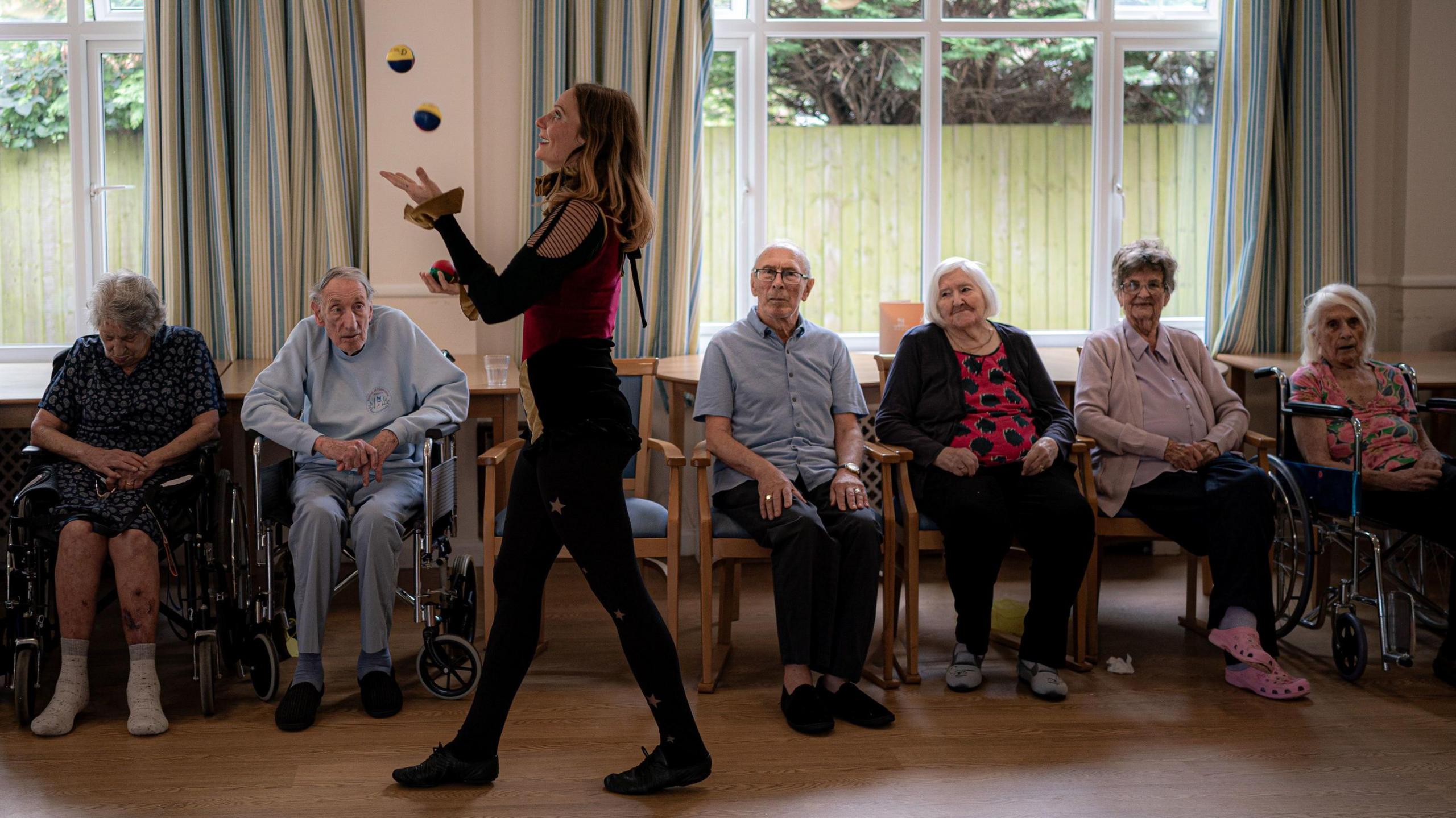 A woman juggling three multi-coloured balls in front of an audience of care home residents.