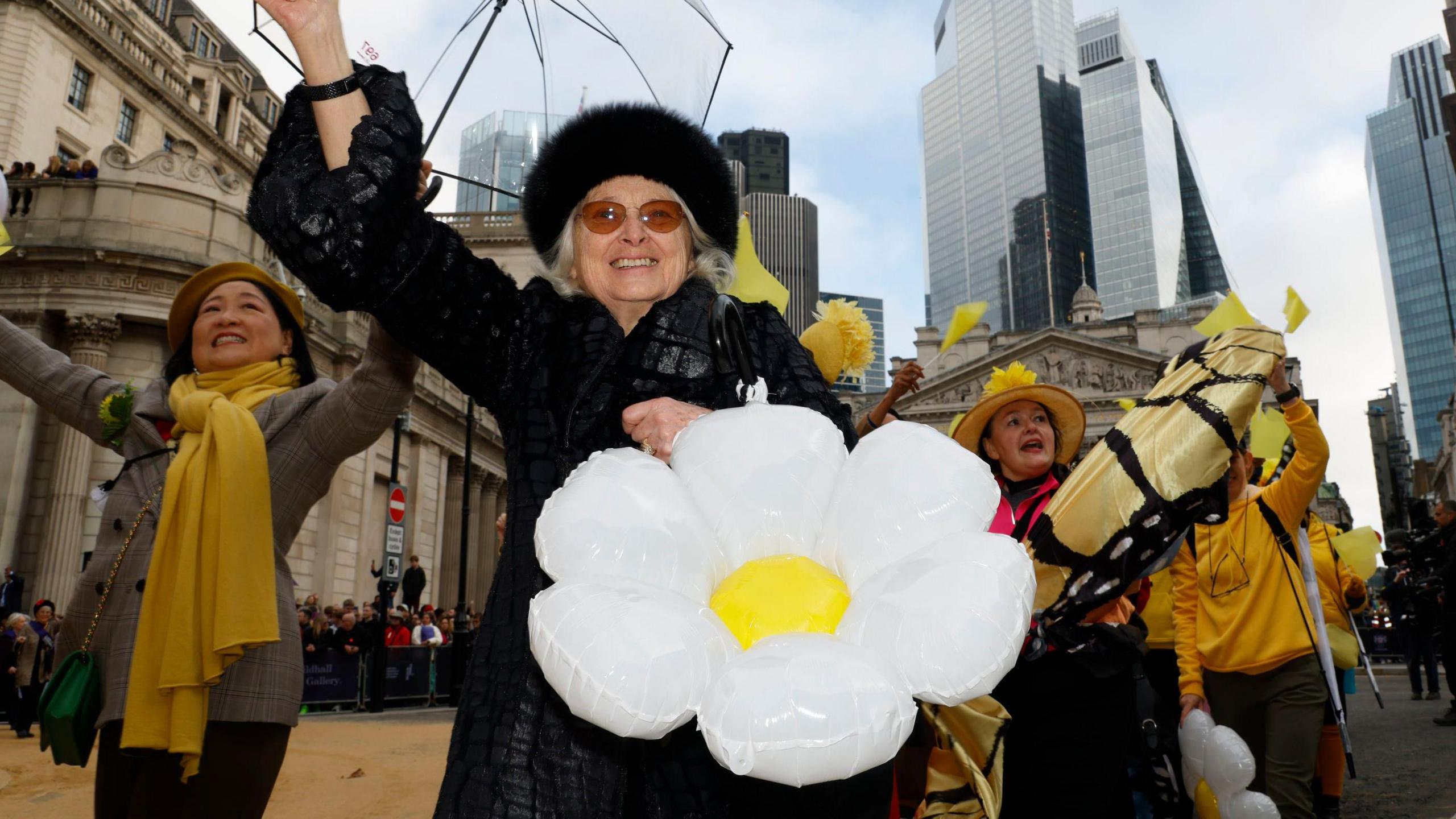 An older woman in a black coat and hat is holding a giant inflatable daisy - she is smiling and waving to the crowds. 