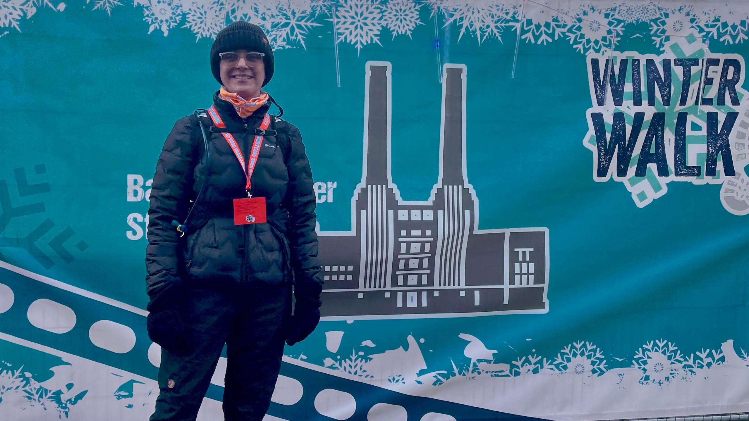 A woman in a black beanie, down jacket, gloves and sunglasses. She is standing in front of a teal-coloured banner.