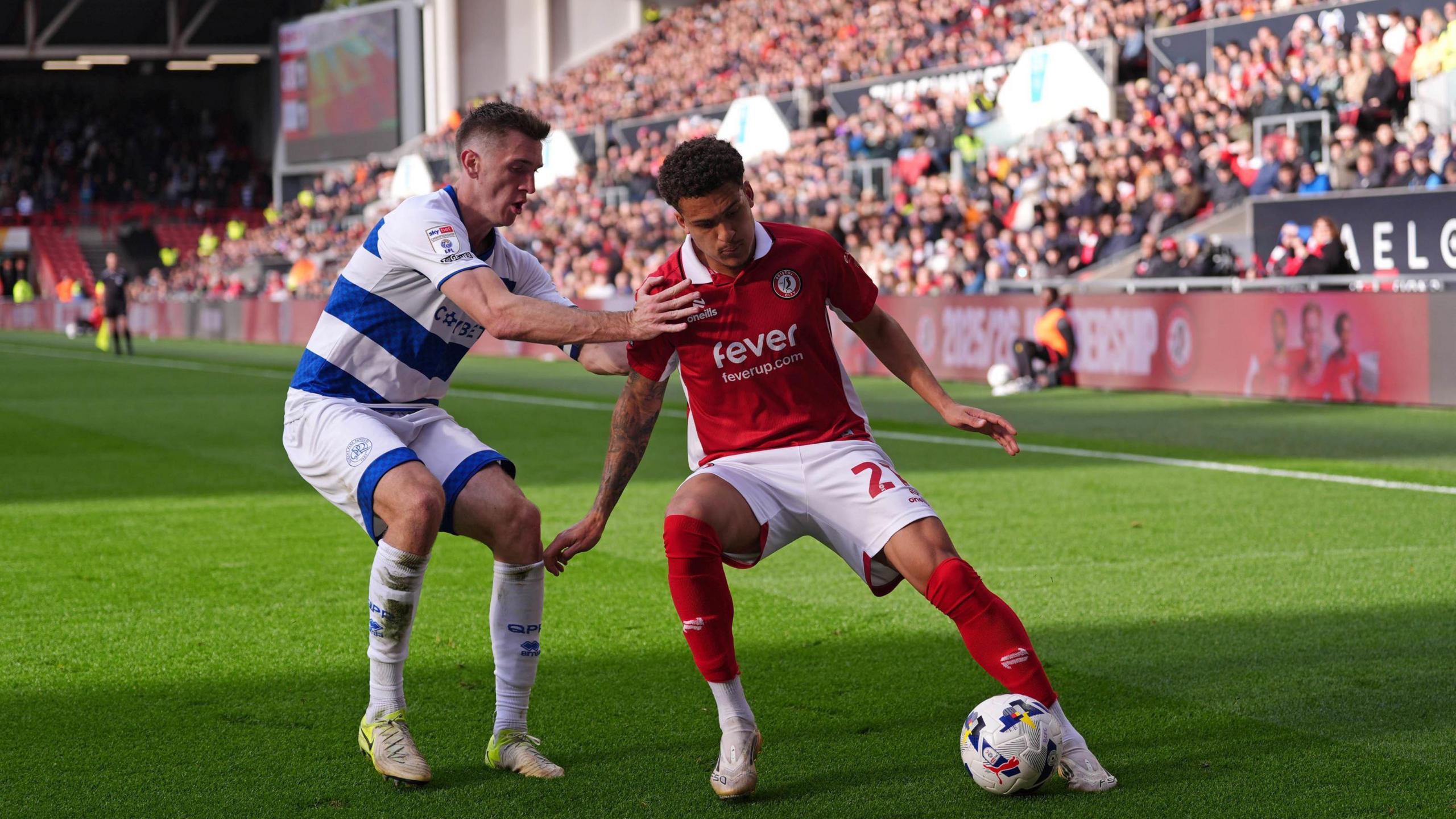 Two players, one from Bristol City and one from Queens Park Rangers, contest for the ball during the match at Ashton Gate. In the background a large grandstand full of spectators is visible