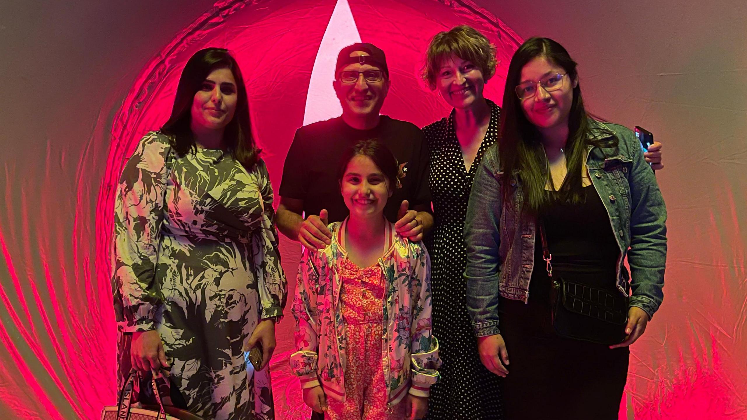 A girl is stands in front of four adults inside a grey, inflatable structure with a bright red light behind them.