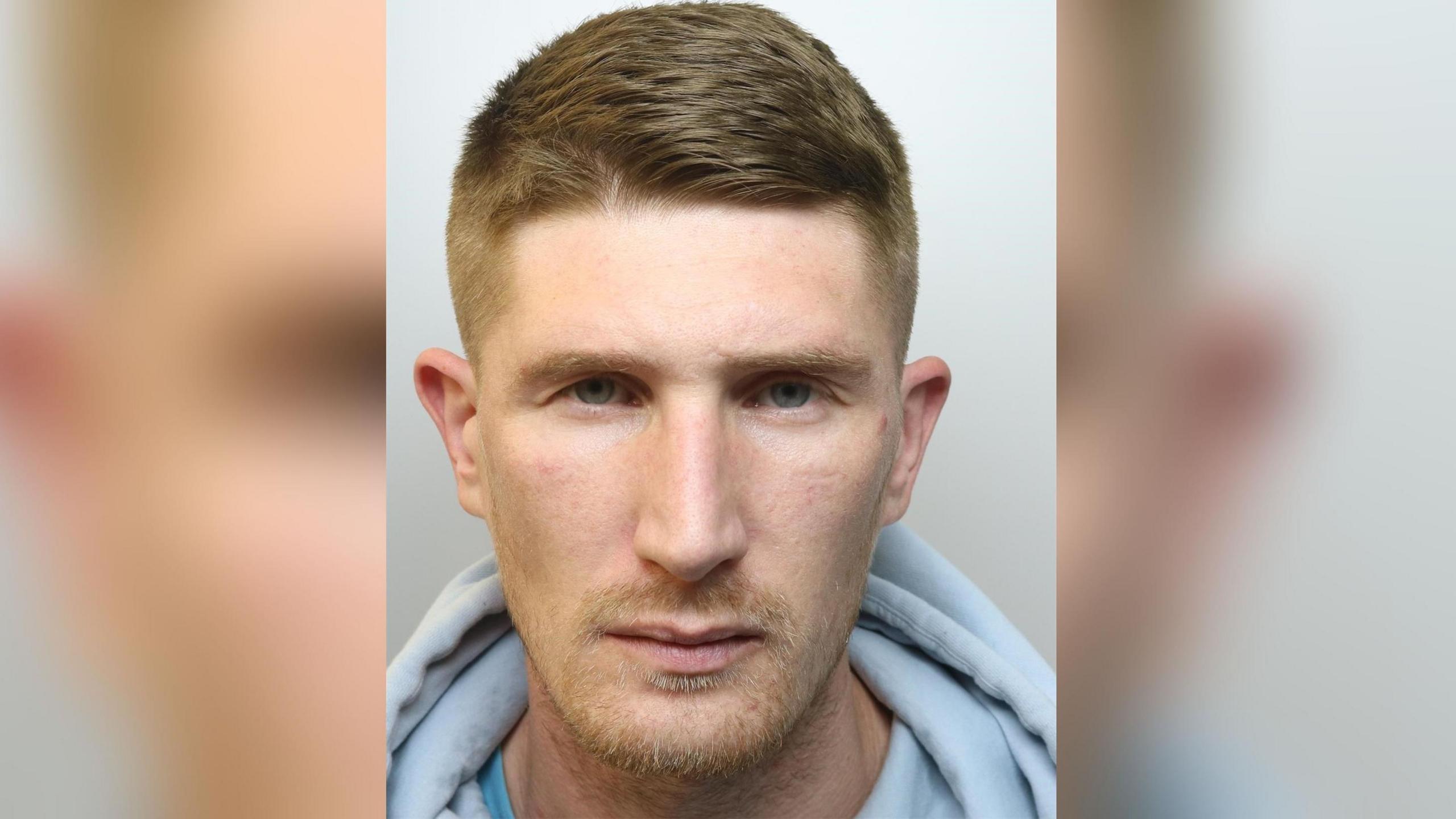 A police mugshot of Jack Cregan, who has short brown hair, blue eyes and some stubble and is wearing a grey top. 