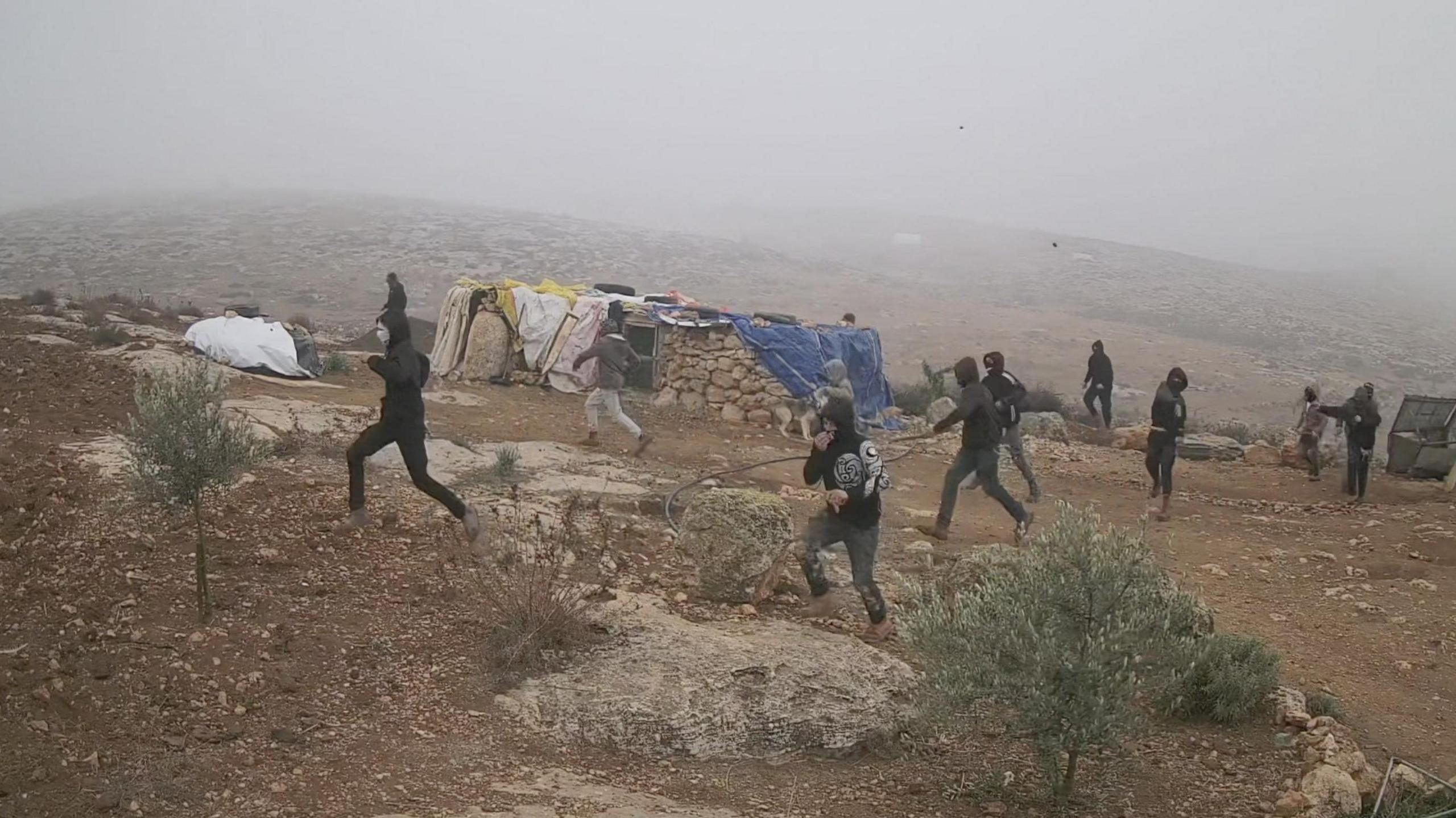 About a dozen people dressed in black with faces covered by hoods and scarves run in the same direction across dry ground, with a small stone building in the background, Khirbet Susiya in the South Hebron Hills, 21 December 2024