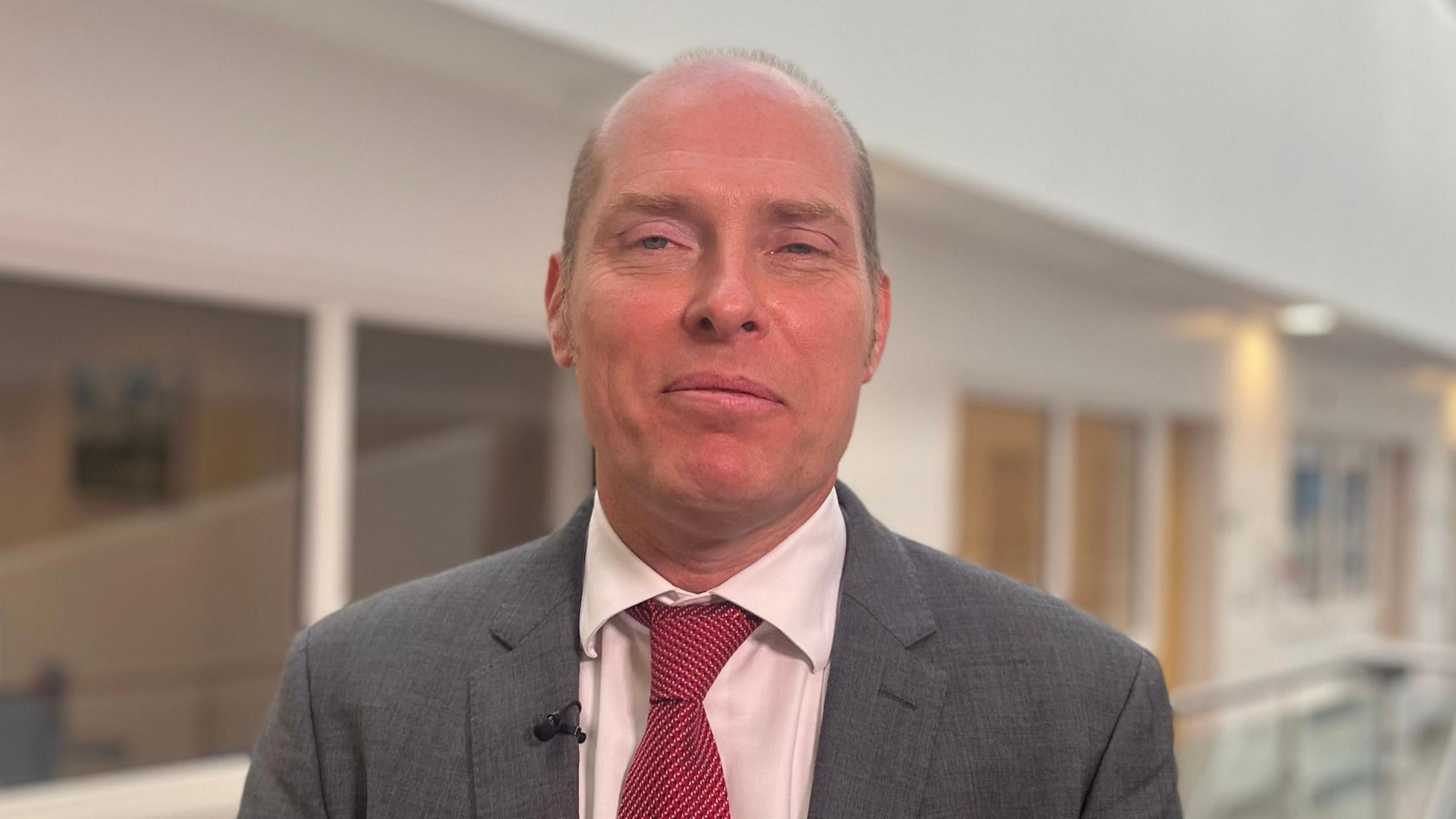 A man, with balding grey hair, smiling at the camera. He is wearing a white shirt, red tie and grey suit. 