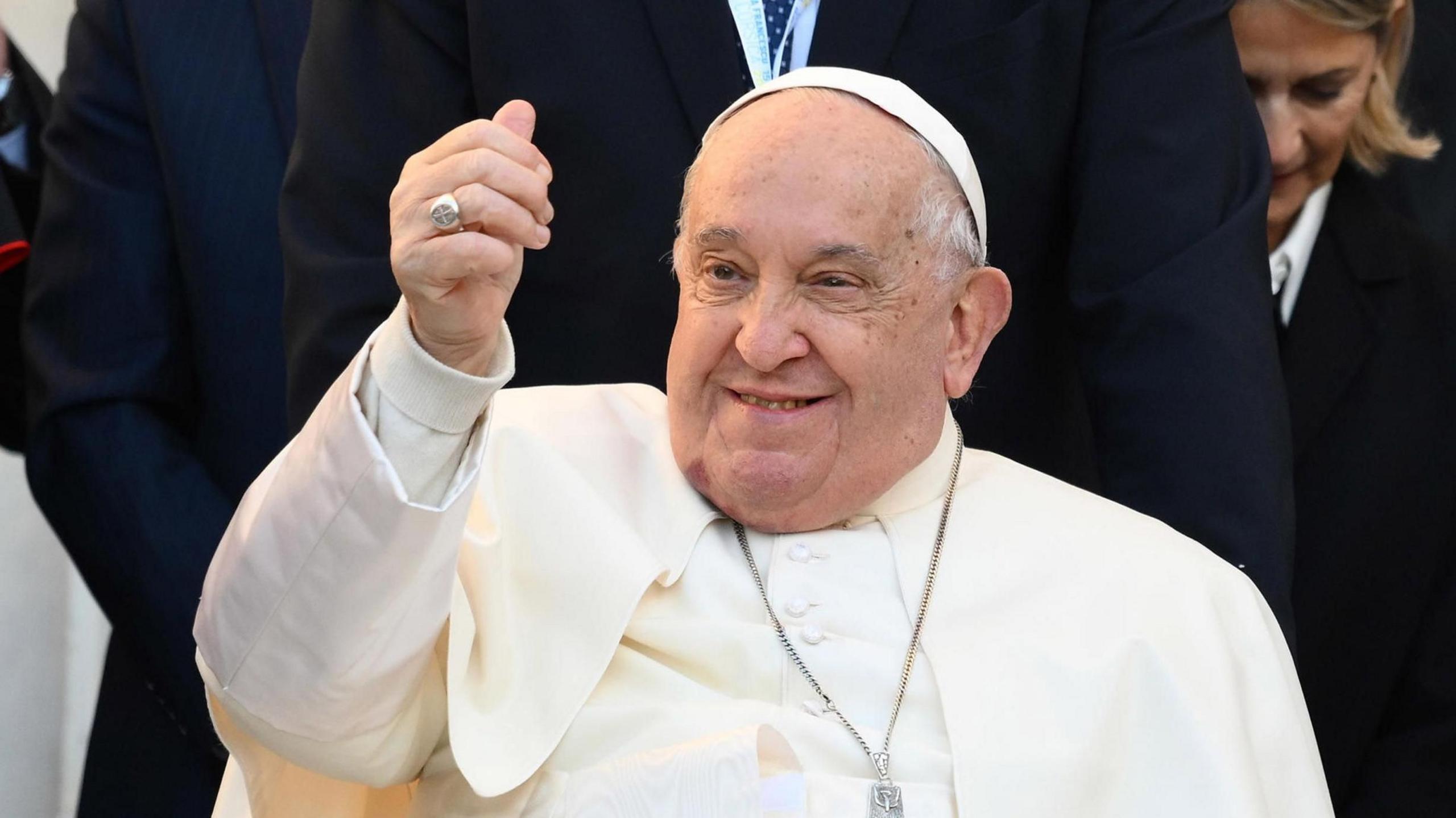 Pope Francis gestures and smiles to the crowds during a papal visit to Corsica in December 2024. He is wearing white robes, a white cap and a crucifix.