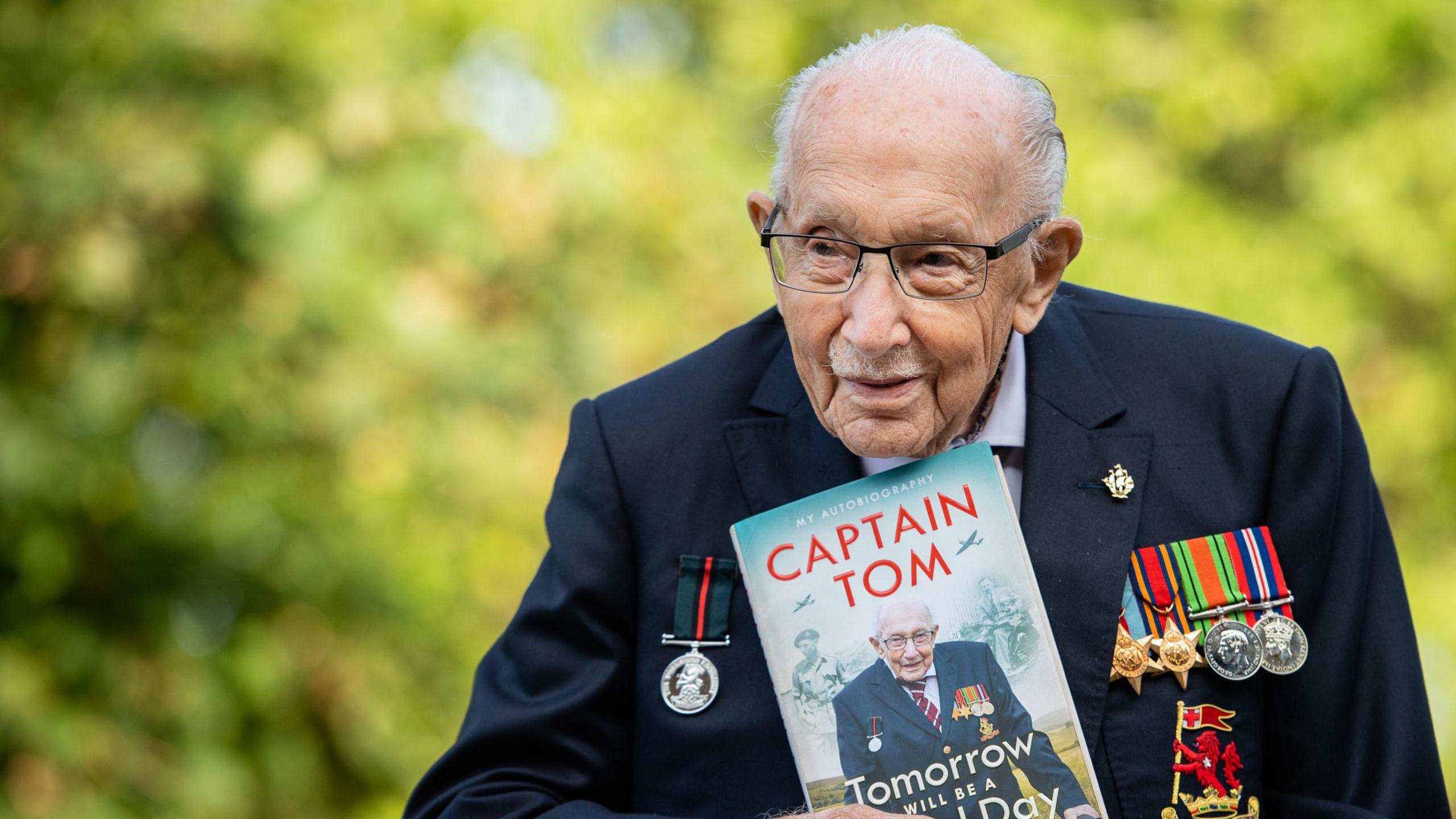 Captain Sir Tom Moore wearing his military medals holding a copy of his autobiography. He has receding white hair, a navy blazer, and there are trees in the background