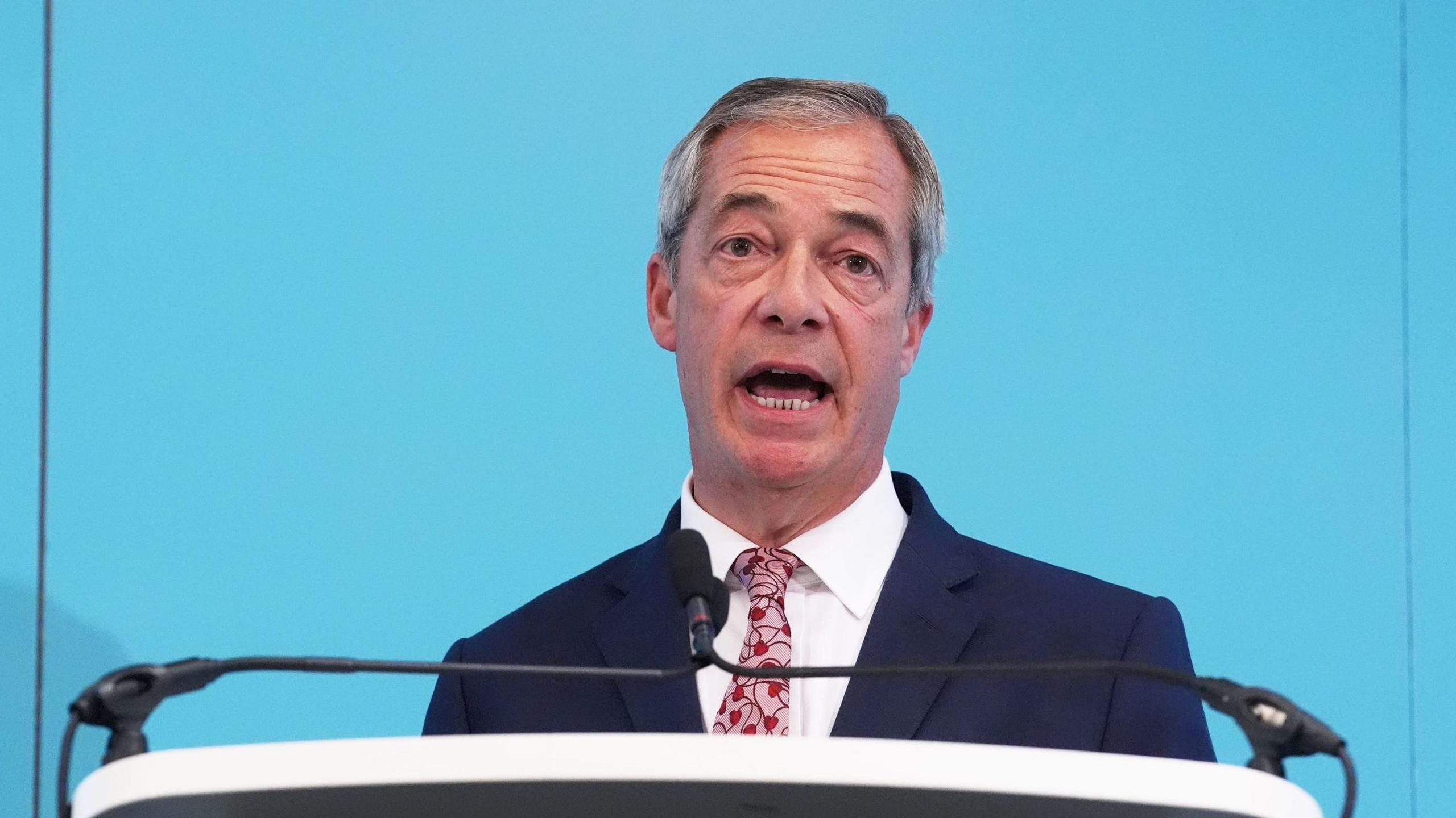 Reform UK leader Nigel Farage stands at a lectern delivering a speech. He wears a white shirt with a bright pink and red tie and a navy suit jacket. He has grey cropped hair.