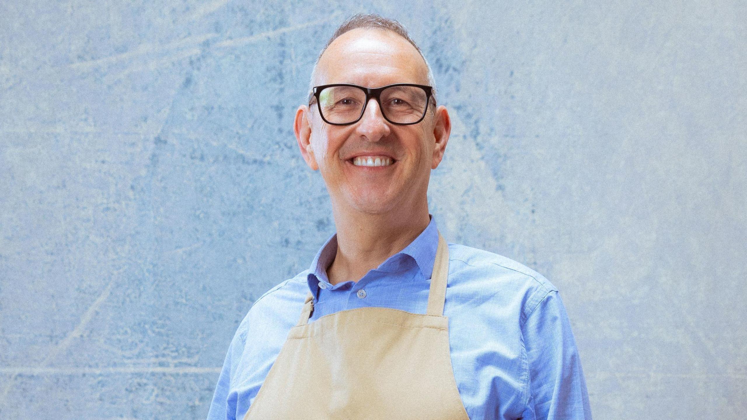 The image shows a smiling man wearing black glasses and a light blue shirt with a beige apron on top. He has short grey hair and the background behind him is patchy and blue.