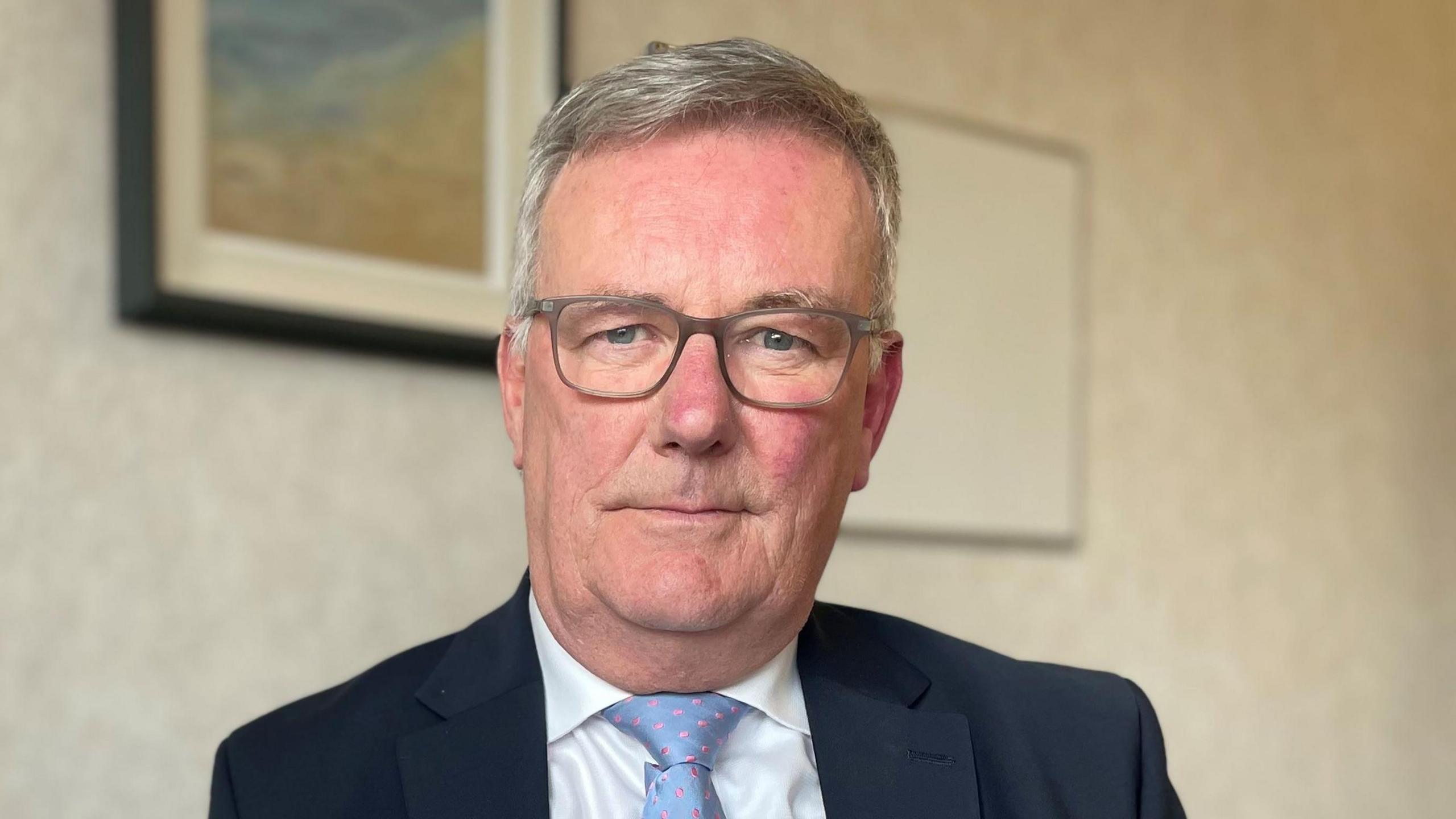 Mike Nesbitt sitting in his office at Stormont. He is wearing a dark blue blazer, a white shirt and a light blue tie. He is wearing glasses and looking directly into the camera.