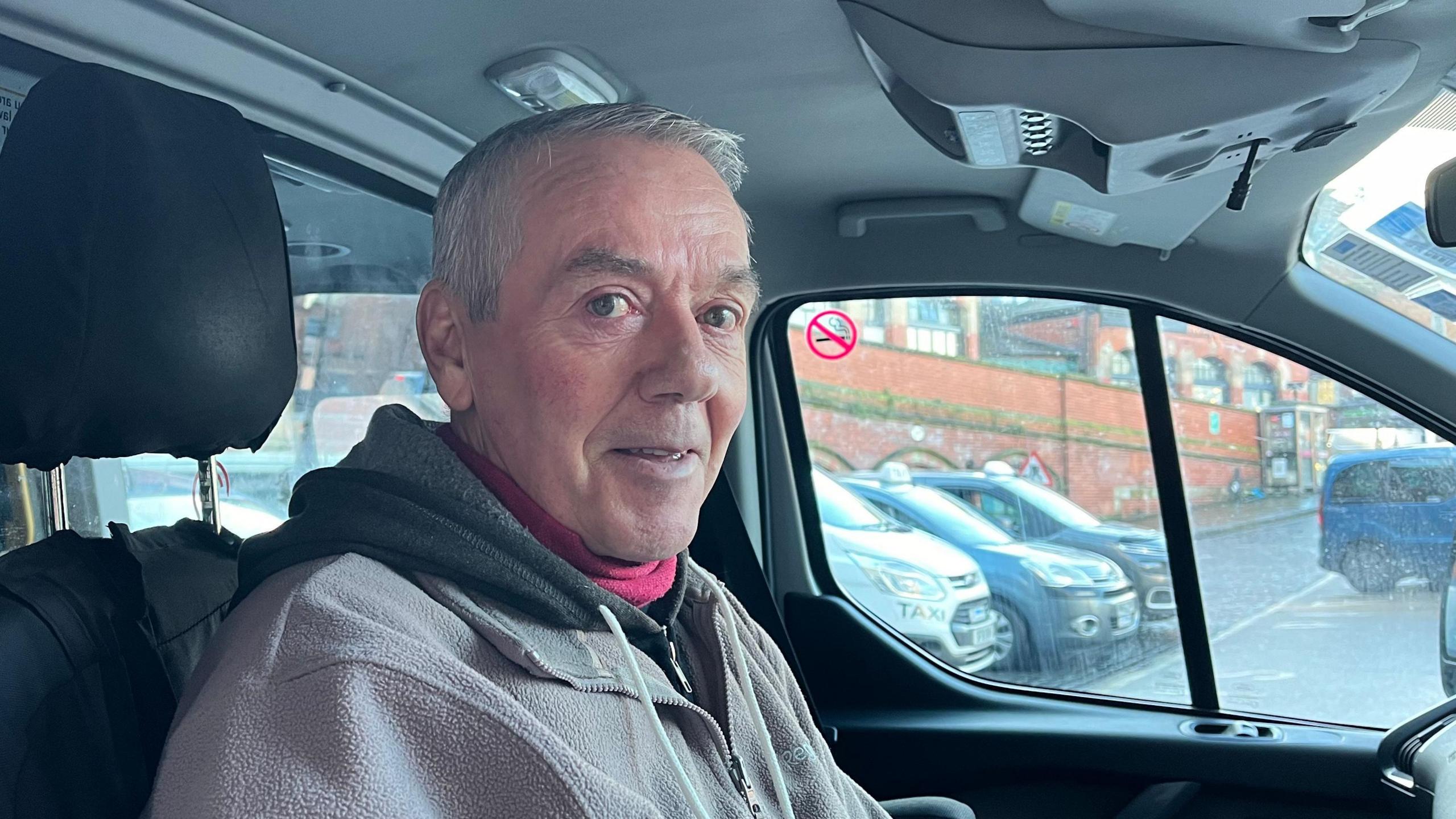 Richard sat in the driving seat of his taxi, he's sideways on to the camera but turning to look at it. Out of the passenger window you can see he's parked up at Shrewsbury railway station, with more taxis visible in the distance. 