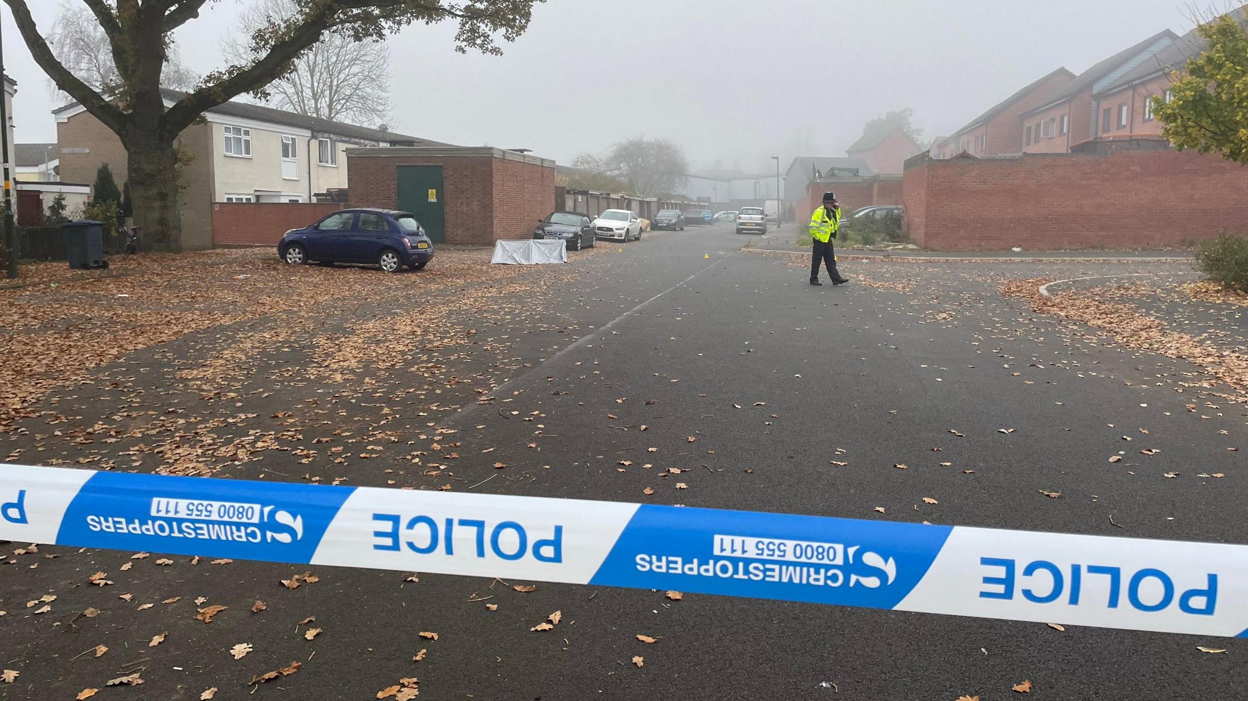 A residential street with a blue and white police cordon. A police officer can be seen walking across the street.