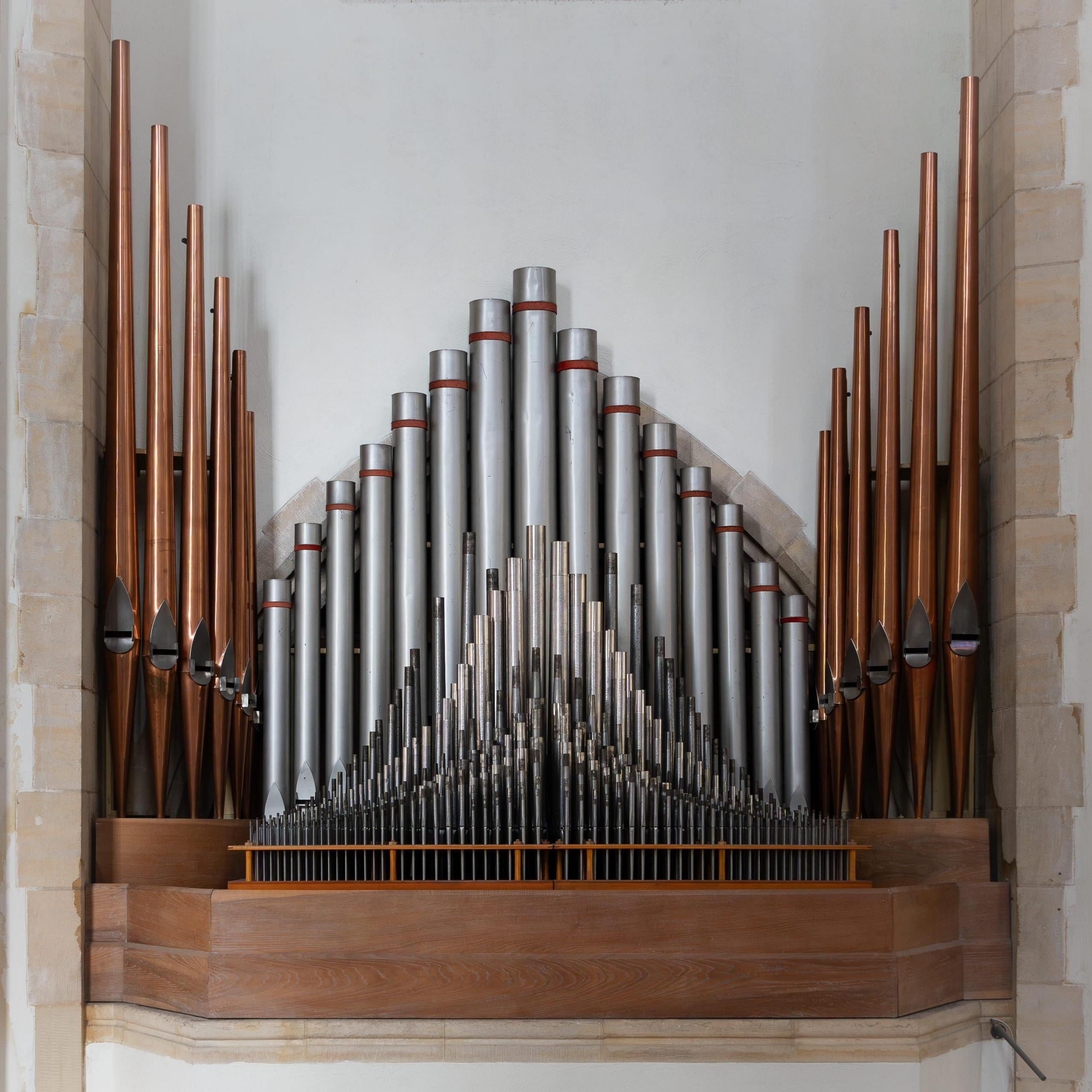 Dozens of organ pipes in varying sizes, silver and bronze in colour. The pipes are set into a white wall with stone brick trim.