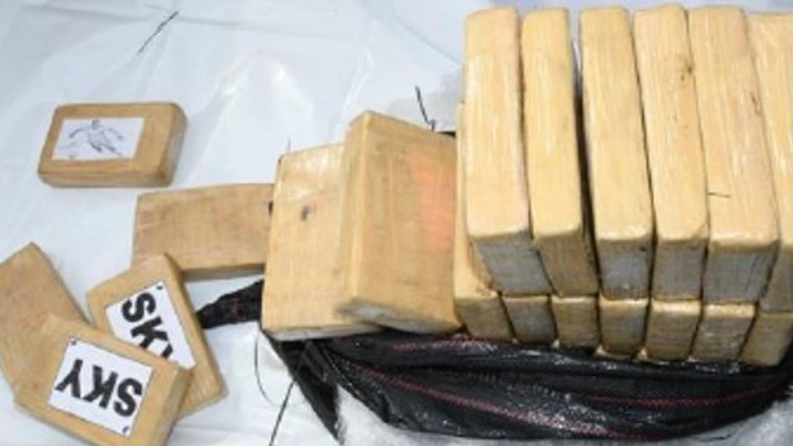 Light brown blocks with the word 'sky' on them. There is a two-storey pile of the blocks, which are filled with cocaine, inside of a black bag. It is on a white sheet.