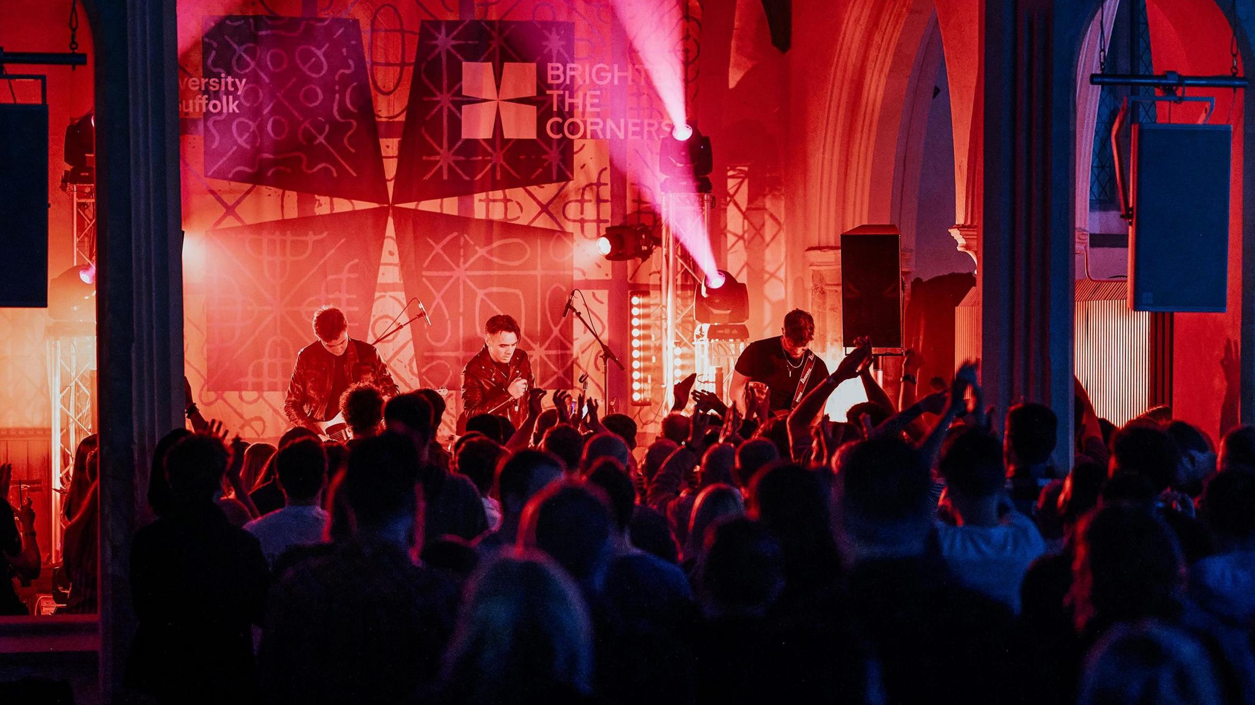 A band performing on stage inside a church. The stage is lit up with a red lighting and three band members. There is a crowd of people facing them. 