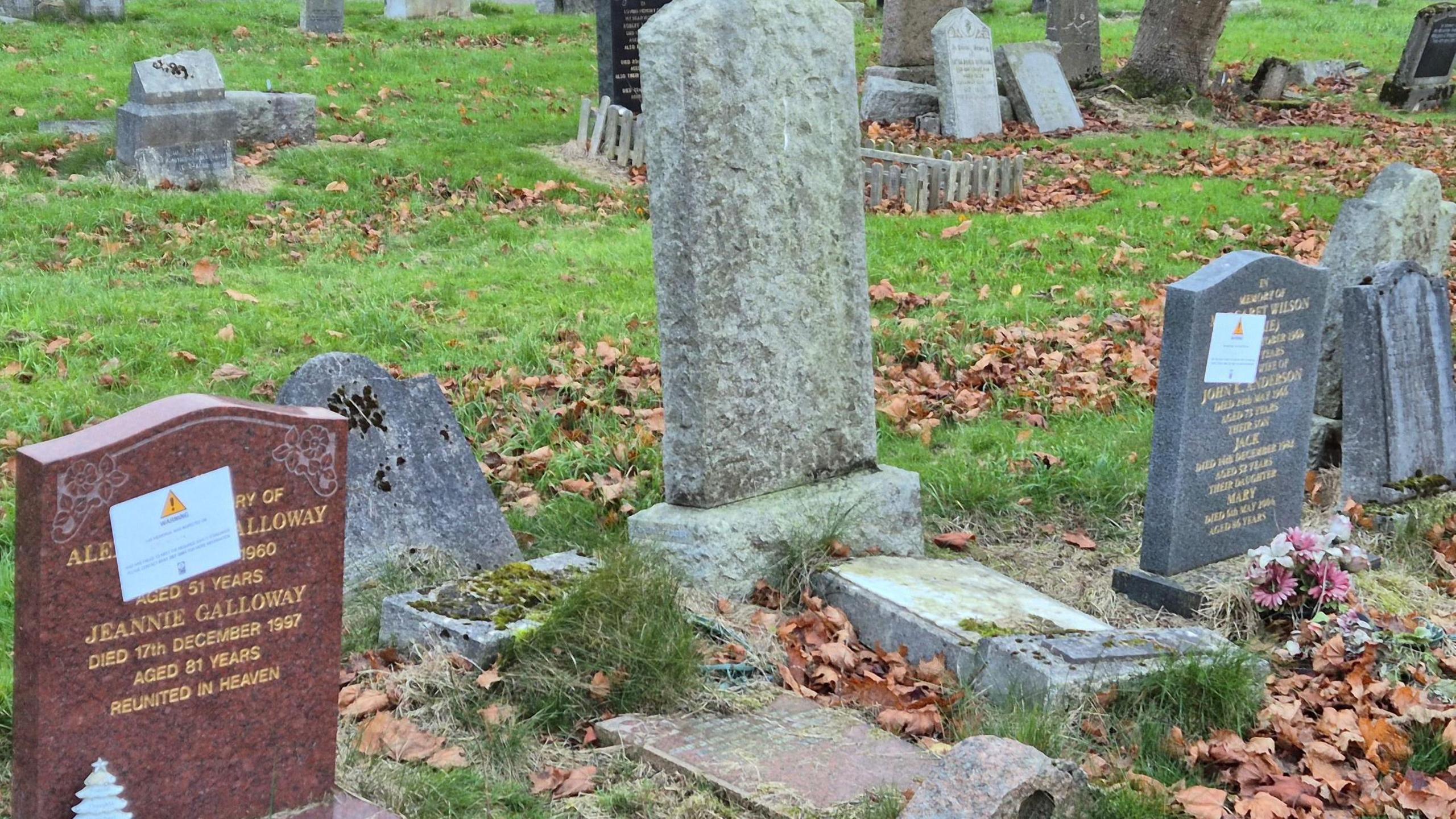 Three graves in Riddrie cemetery - the left hand one is a reddish brown and has a warning sticker applied, while the right hand one is grey and also has a sticker applied. The one in the centre is grey and unlabeled. 