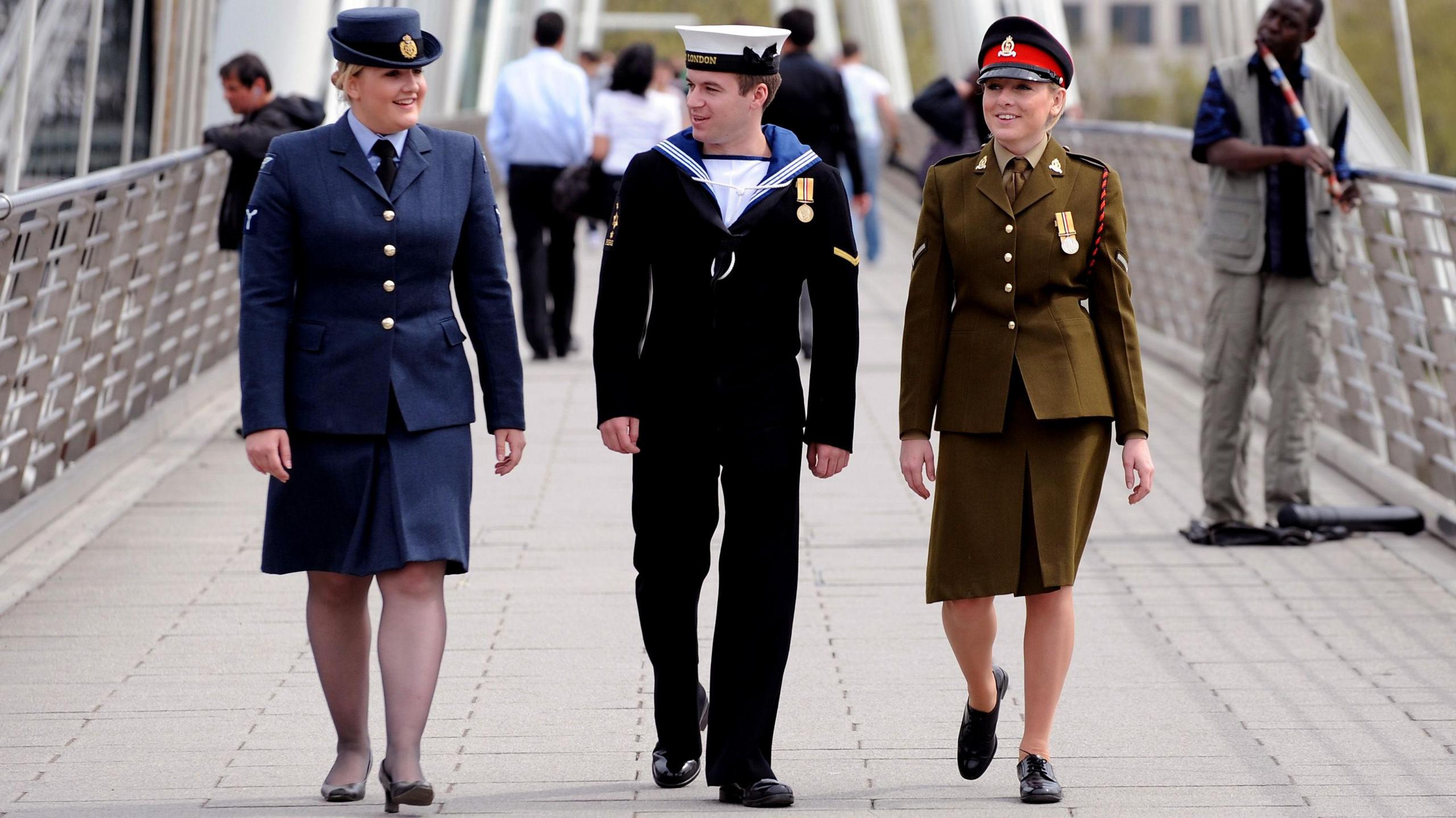 Three uniformed military staff, one man and two women, walk side-by-side across a bridge. One is in a navy blue uniform with a hat and skirt, one is in dark trousers and top with blue collar and white hat, and one is in a brown skirt and buttoned blazer with a hat. 