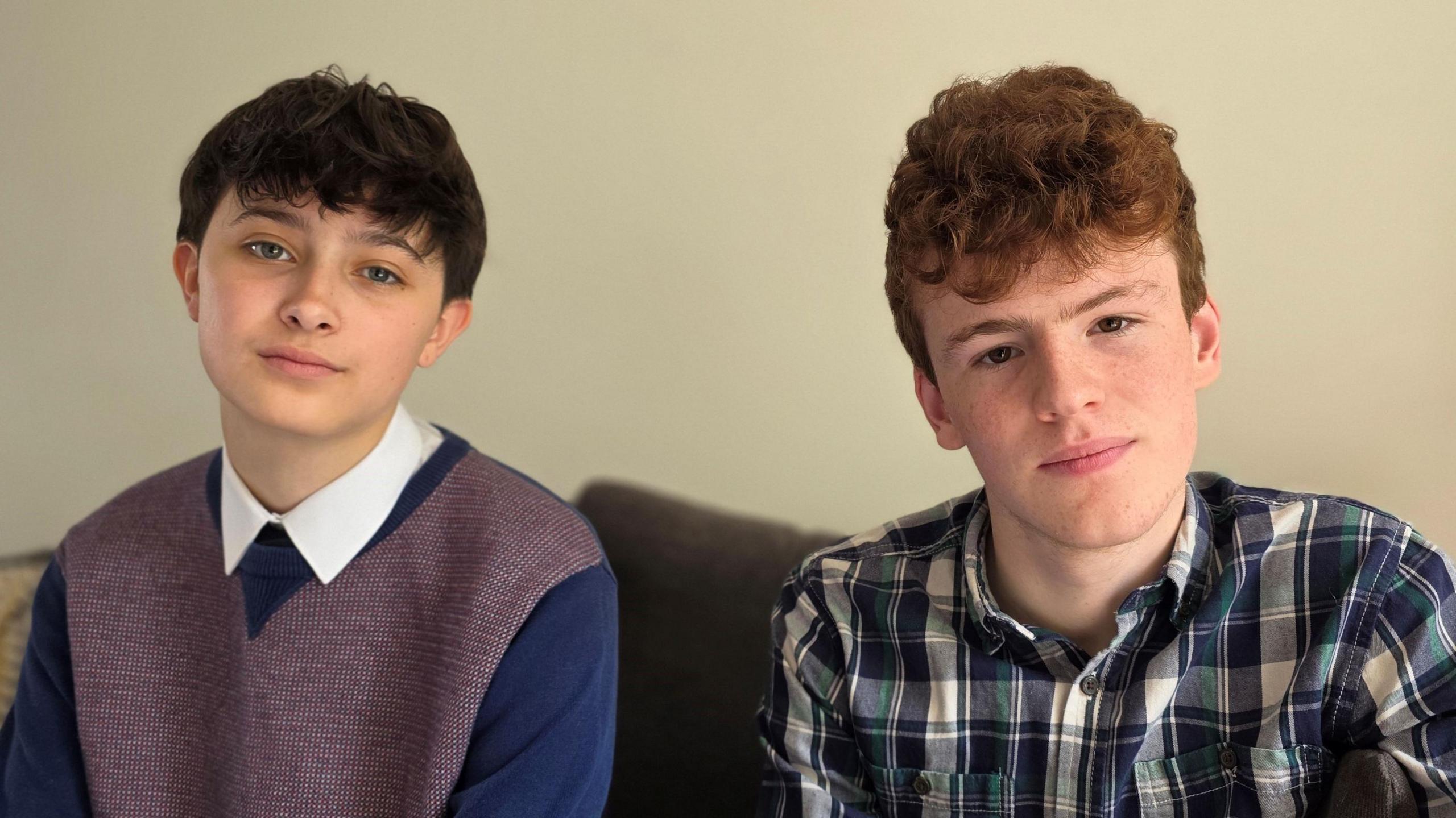 Arlo is on the left side of the image wearing blue sweater over a white-collared shirt. George, on the left, has a blue and white checked shirt. They are sitting on a sofa.
