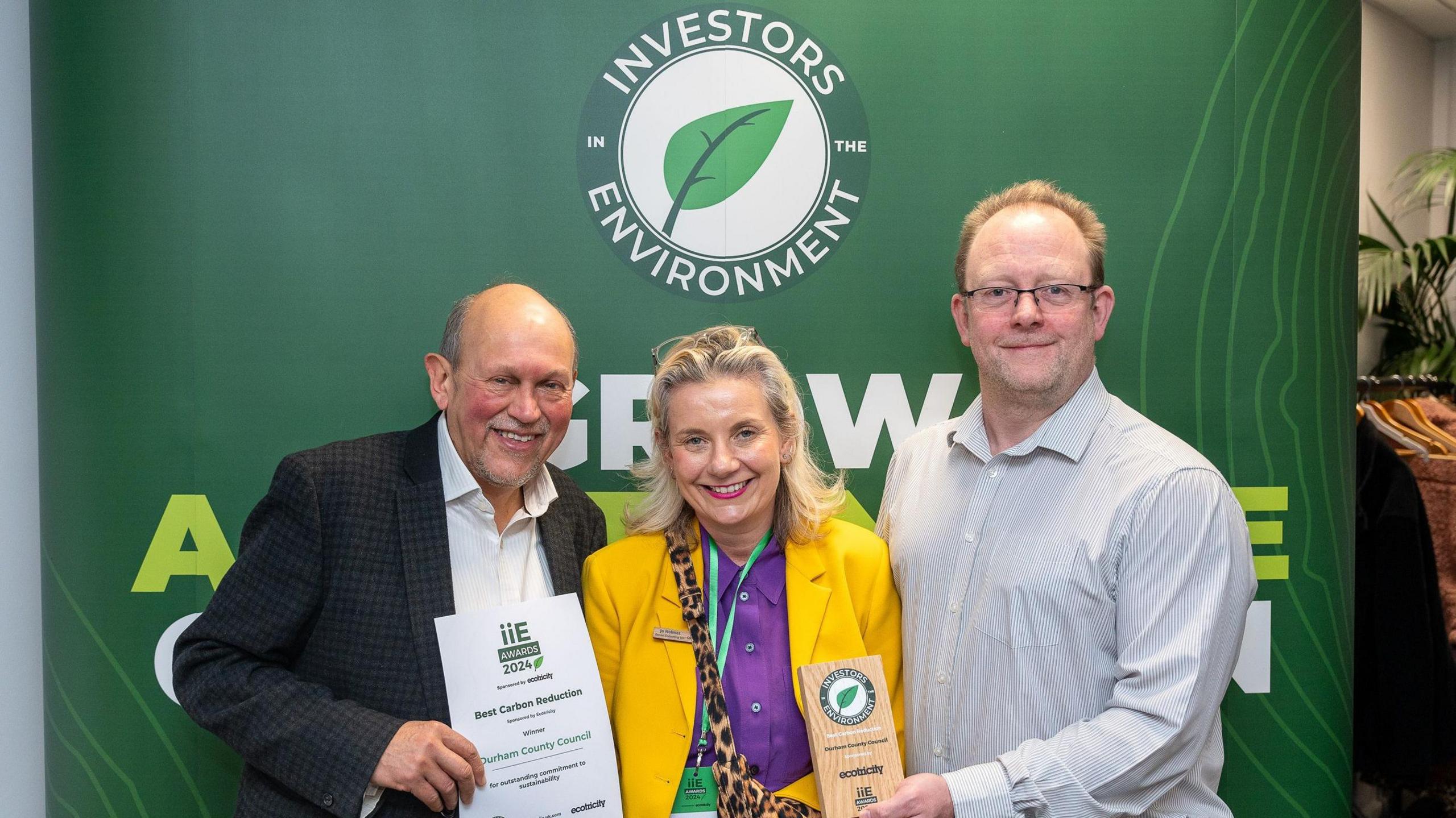 Steve Bhowmick, environment and design manager from Durham County Council; Jo Holmes, manging director of Genee Consulting Ltd; and Stephen Beresford, principal officer climate strategy and policy at Durham County Council. They are holding their certificate and award. They are standing in front of a banner that reads, Investors in the Environment.