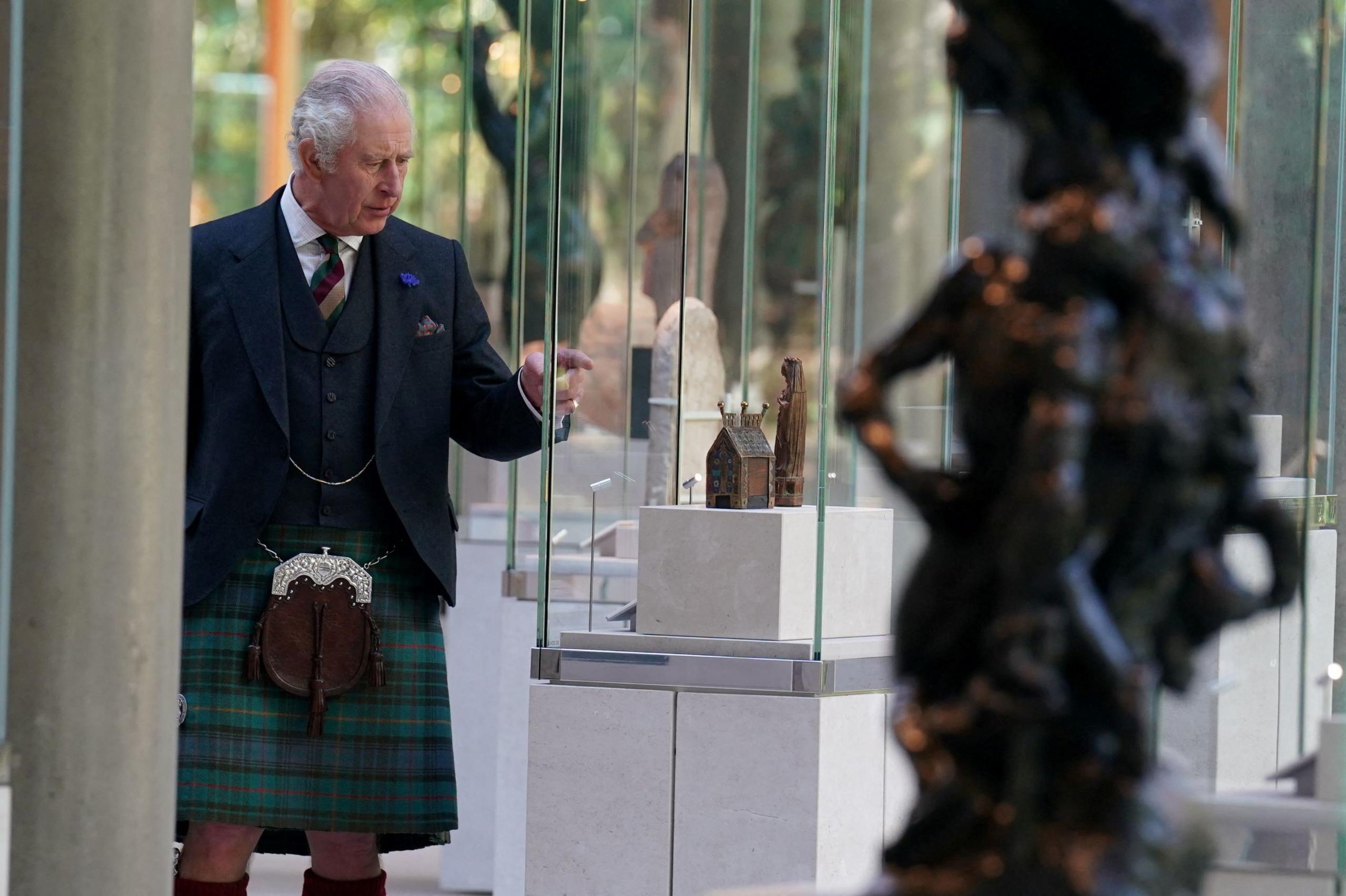 King Charles, dressed in a dark green kilt and dark jacket, inspects an exhibit in the Burrell Collection. There is a sculpture in the foreground and we see him behind this.