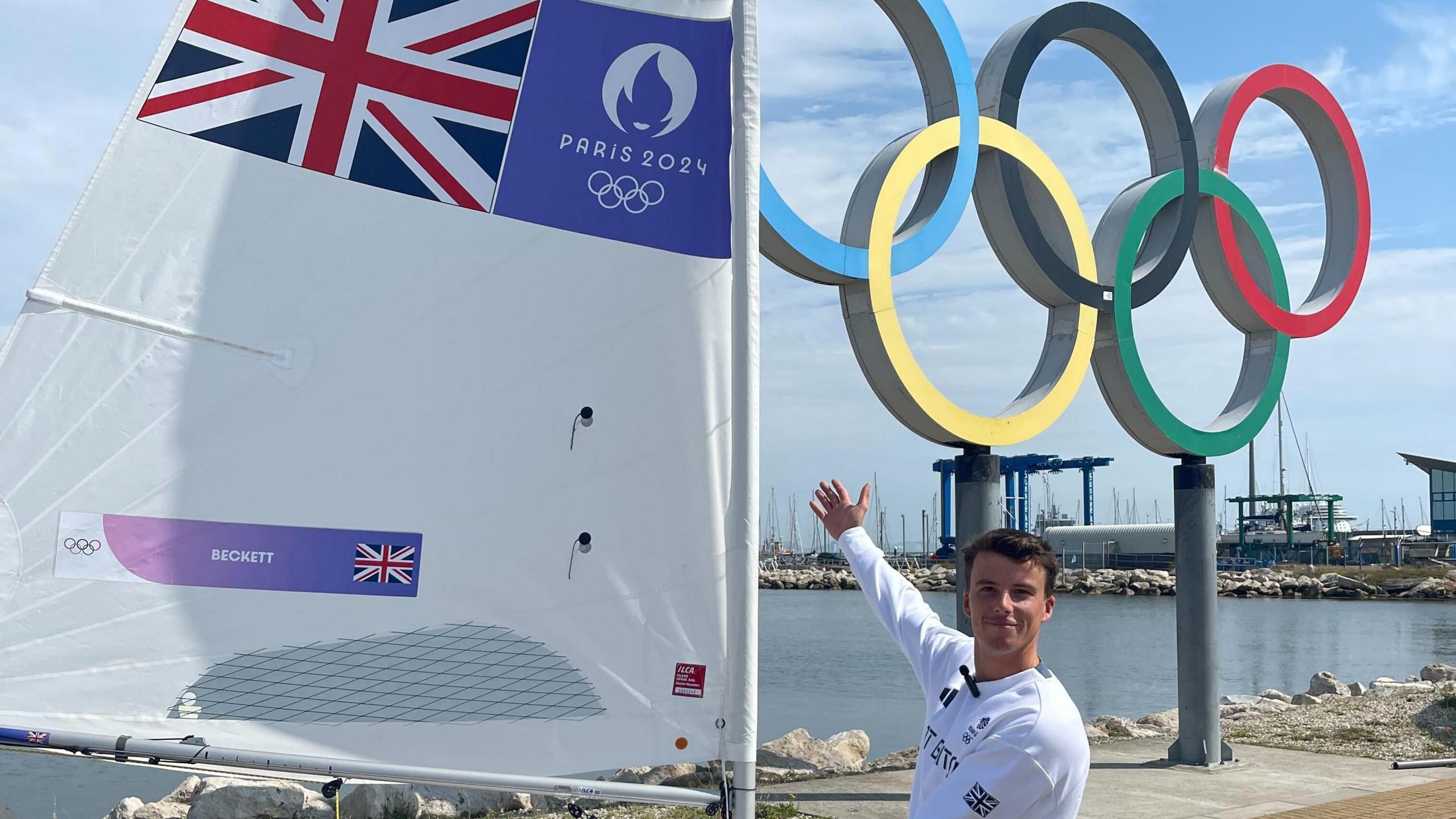 Micky Beckett, wearing a white Team GB t-shirt, stood next to the boat he had used at this years olympics. The olympic rings are on a plinth behind him.