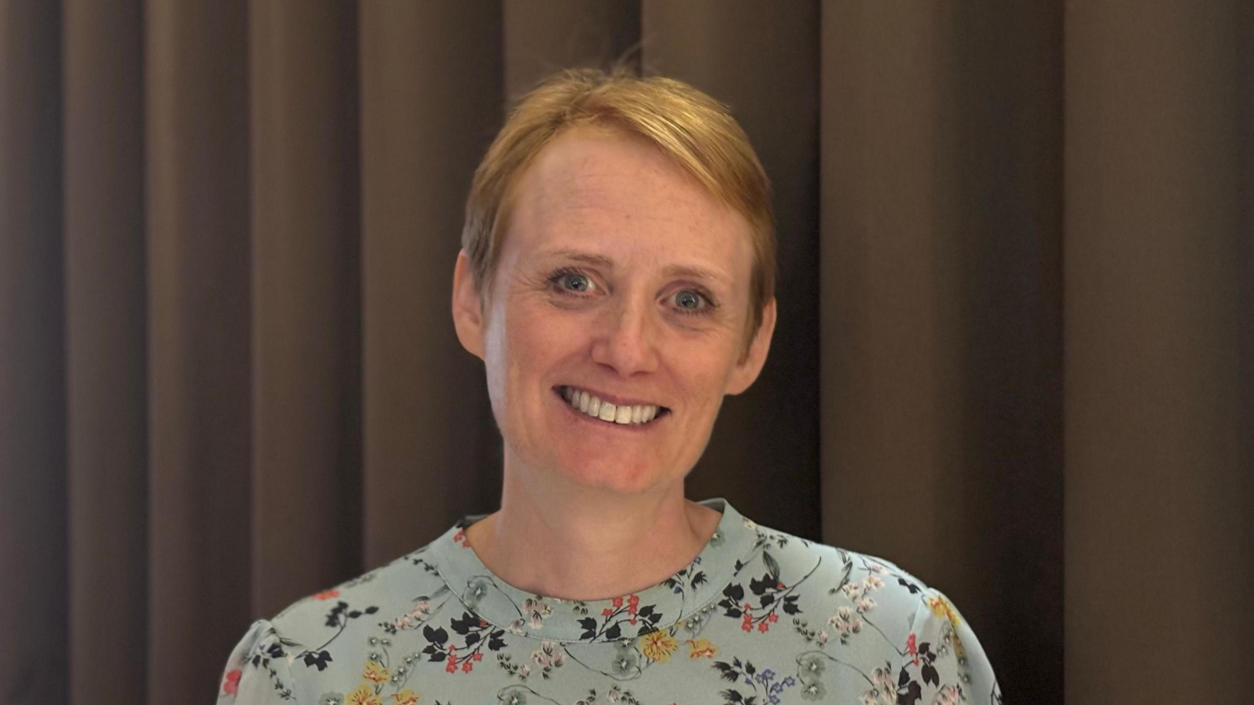 Jane Burns photographed wearing a light blue top with a floral pattern. She has short ginger hair. 