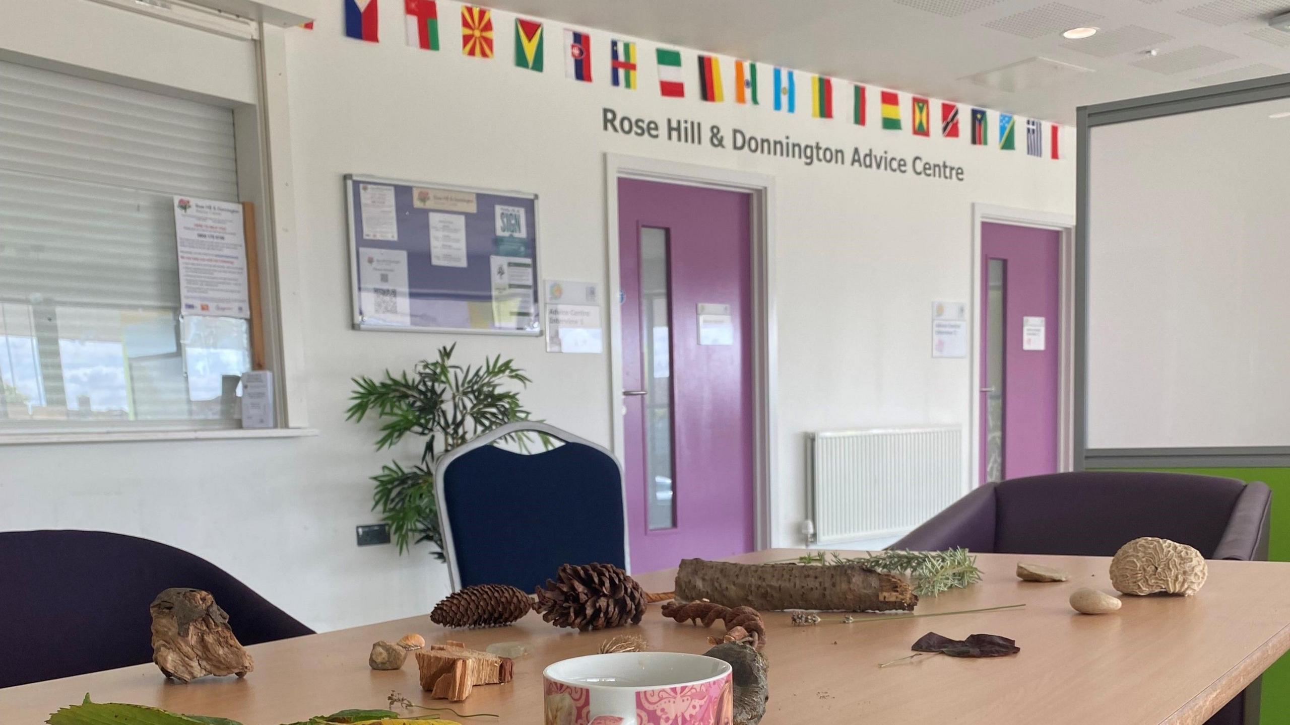 A table in a meeting room covered with items from the natural world such as acorns, twigs and rocks. There is also a coffee cup on it. Chairs surround the table. A sign above the door reads Rose Hill and Donnington Advice Centre. The flags of multiple countries are placed above it.