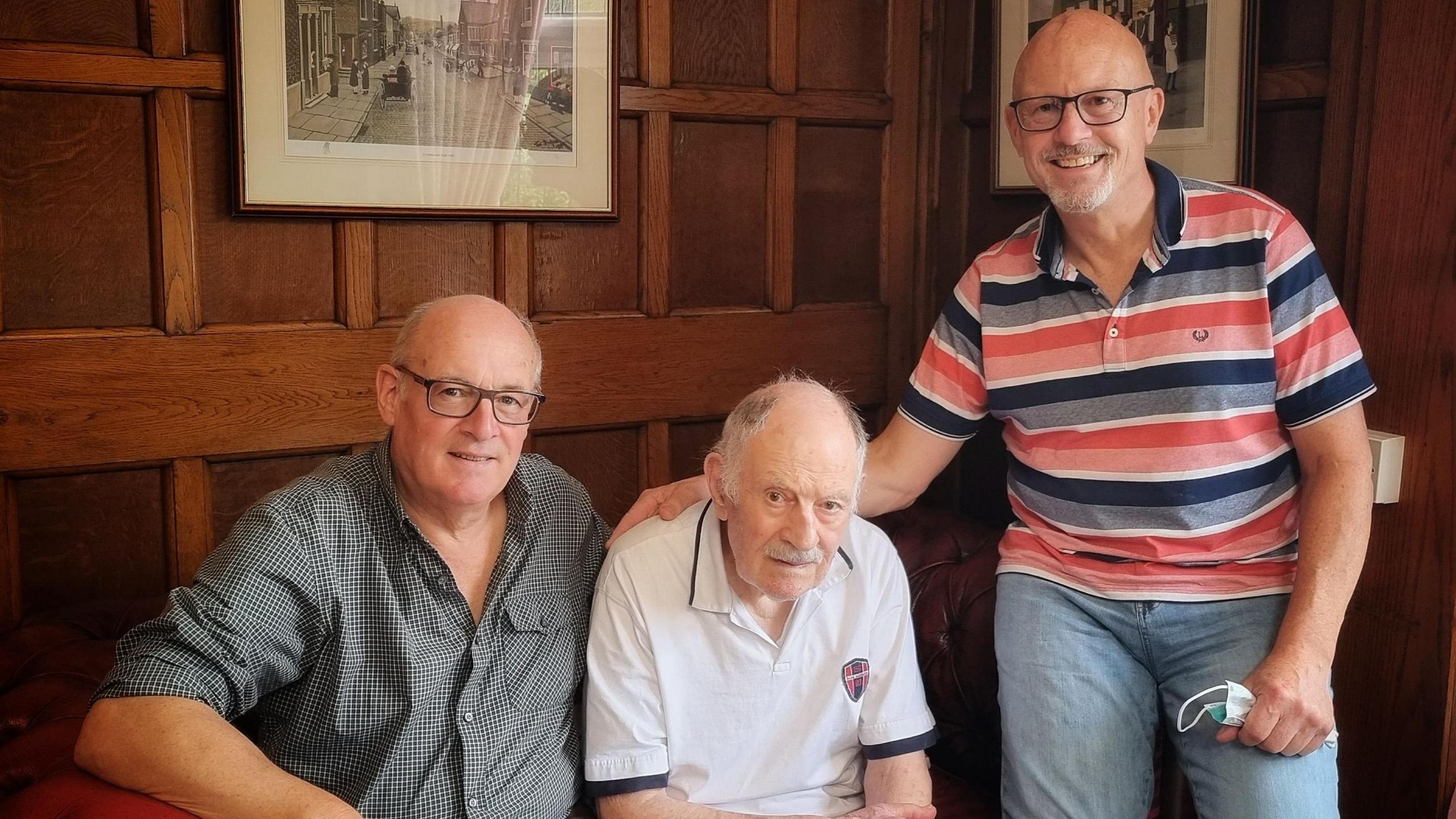 Left to right: Martin Taylor wearing glasses and a black and white checked shirt, Edward Taylor with a white moustache and white hair wearing a white polo shirt both sitting on a sofa and Paul Taylor smiling and standing with his arm round his dad wearing a red blue and white striped top.