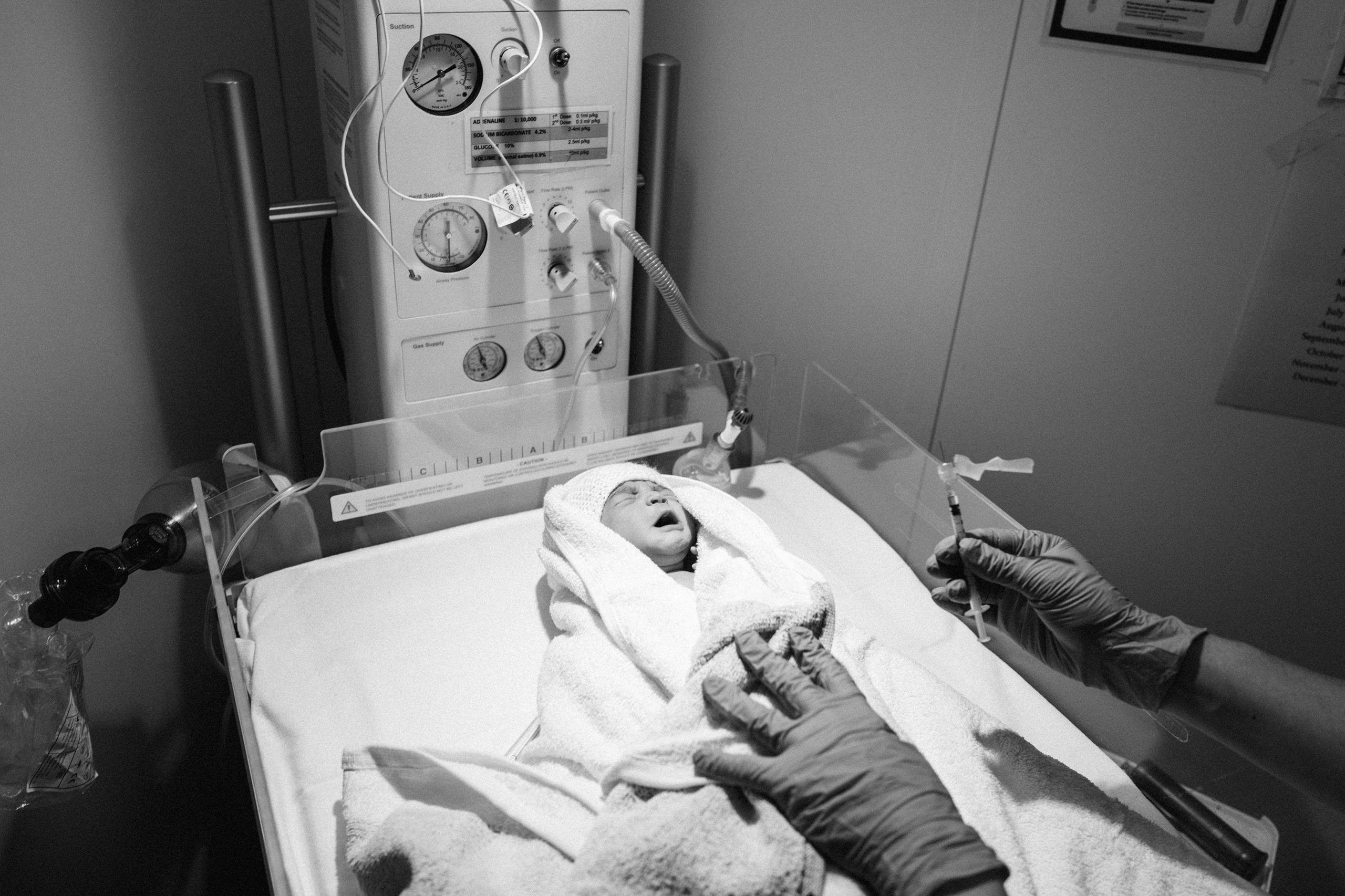 The newborn is seen wrapped in a towel on a hospital bed, receiving a Vitamin K injection from a healthcare worker in gloves. Medical equipment is visible beside the infant.